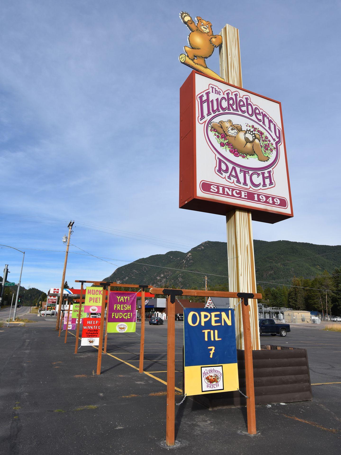 A sign for The Huckleberry Patch in their parking lot. There are animated bears on the sign and it reads "since 1949." Closer to the ground, signs read "open til 7" and "Yay! Fresh Fudge" and "Huck Pie!"