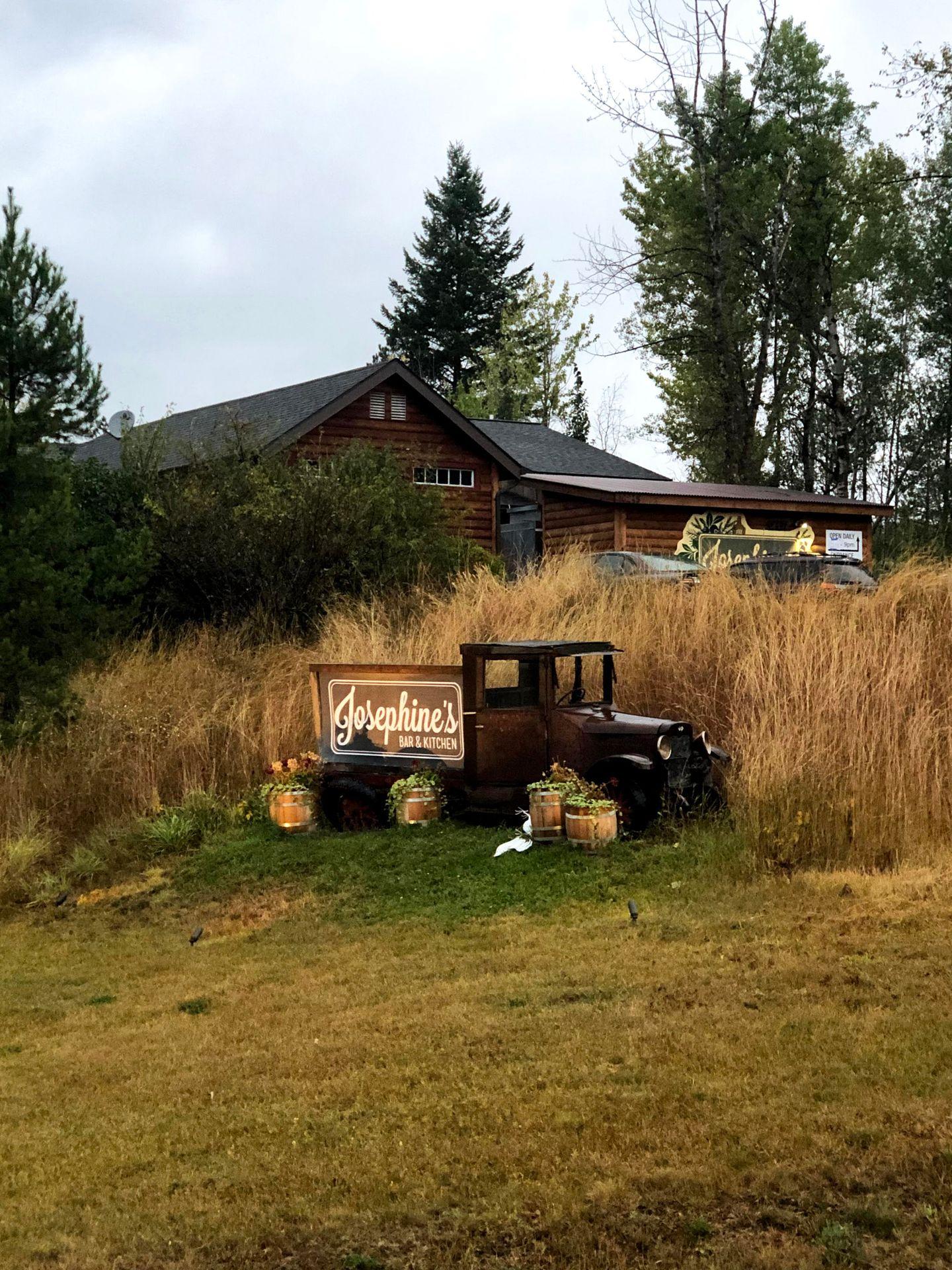 A vintage pick up truck in front of tall grass. There is a sign of Josephine's in the bed of the car.