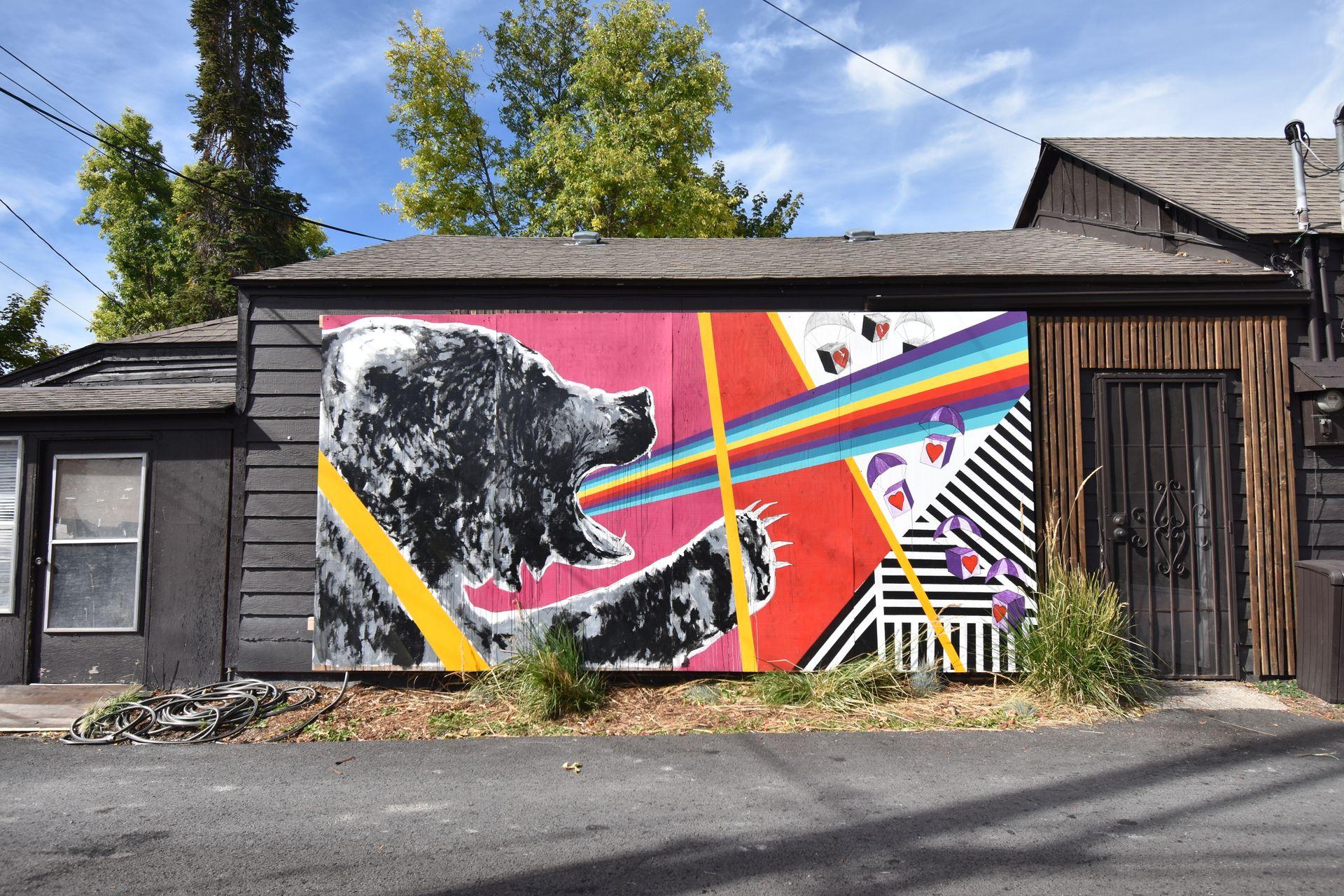 A mural of a bear with a rainbow coming out of its mouth. There are pink, red, yellow and black and white geometric shapes surrounding the bear.