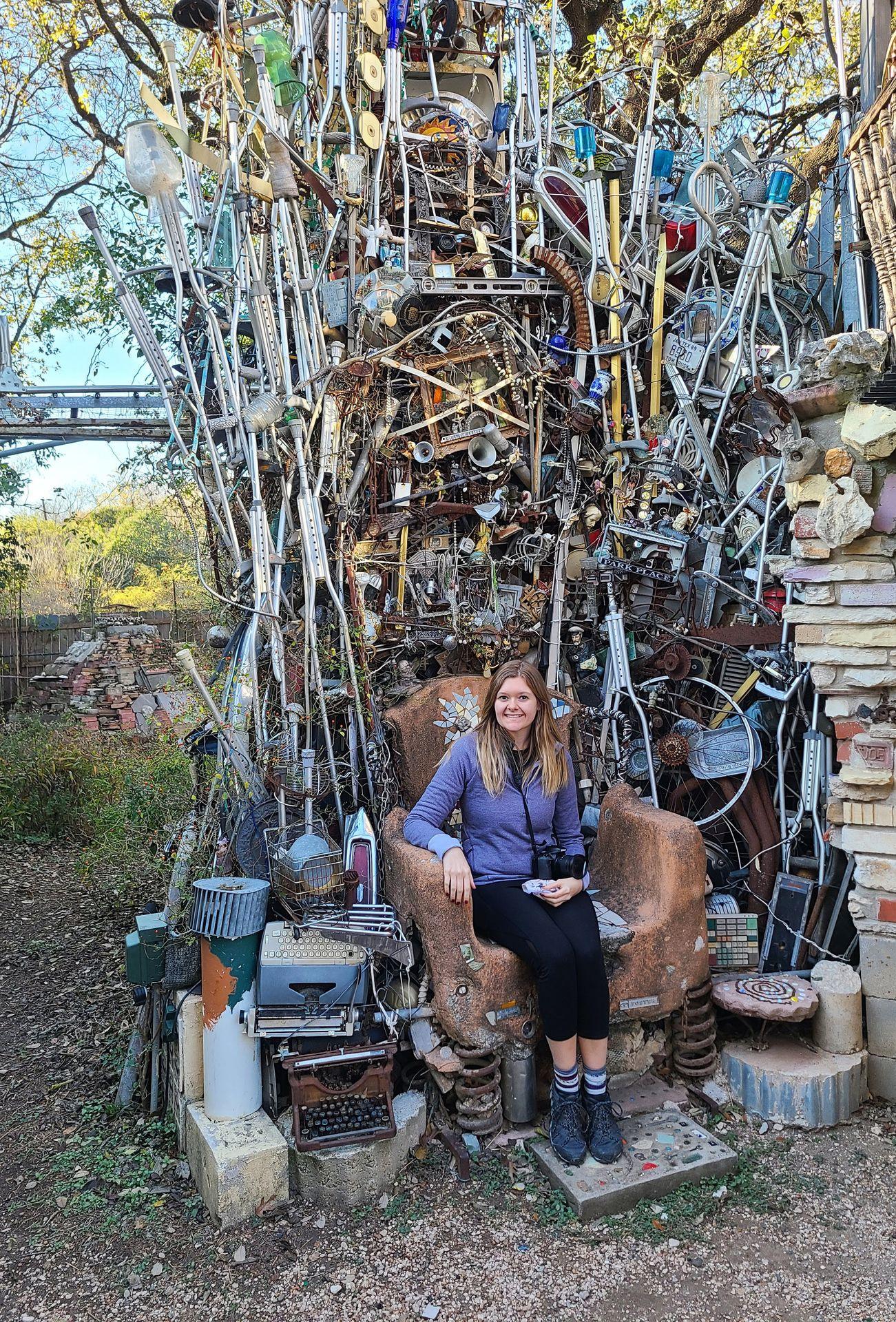 Lydia sitting on a thorne chair with objects built up behind her at the Cathedral of Junk.