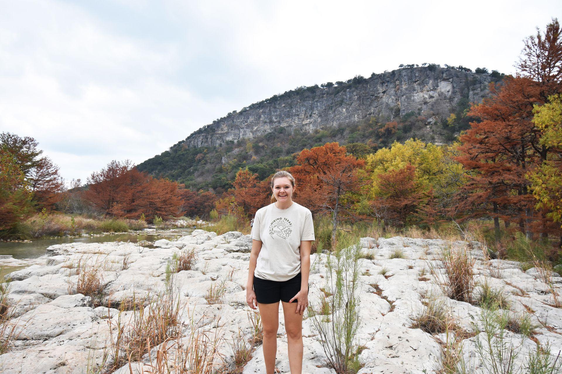 Lydia standing on rocks next to the Frio River with fall foliage in the background.