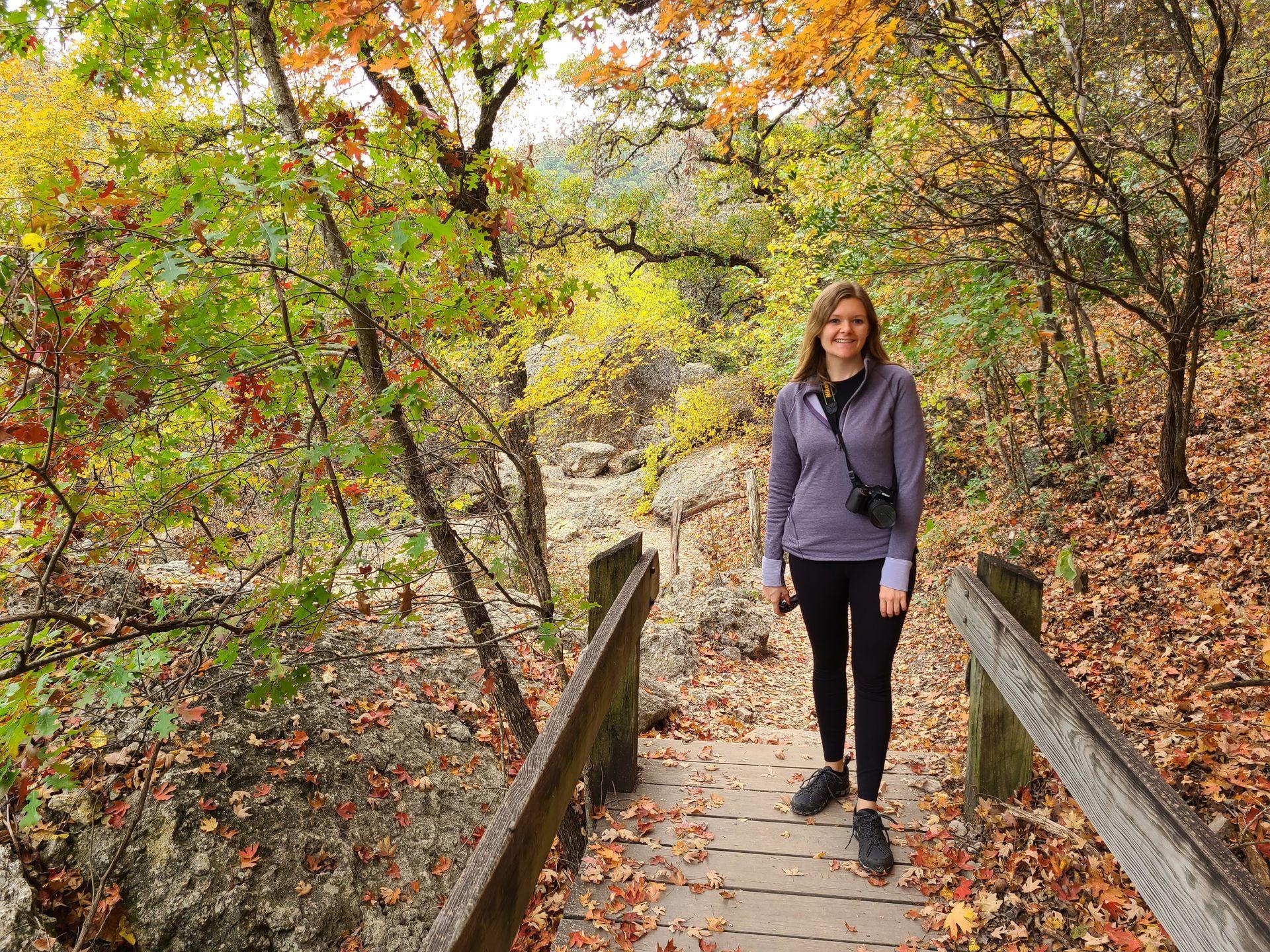 Lydia standing on a bridge with colorufl fall foliage behind her in Lost Maples.