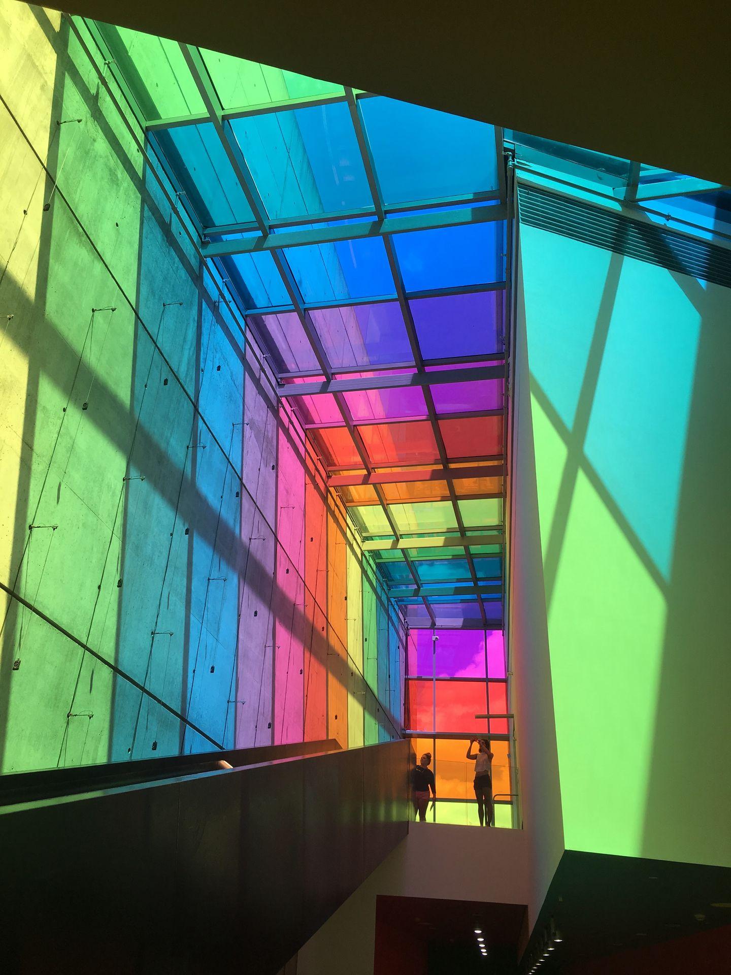 A hallway in the interior of the CAC that is lit to be rainbow colors. There are windows at the top and shadows created by the window panes.