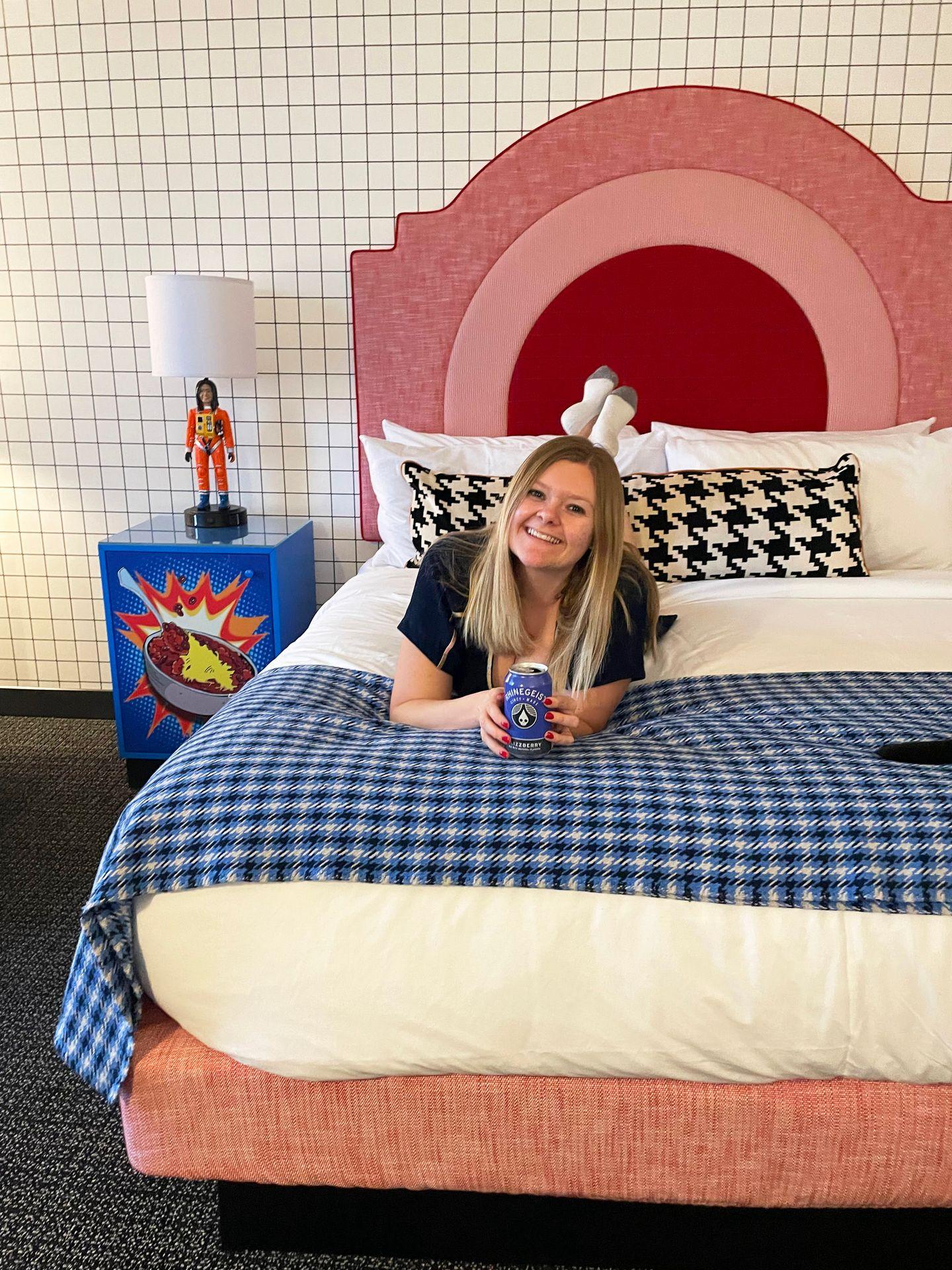 Lydia laying on her stomach in a room in the Graduate Cincinnati hotel. She holds a Rhinegeist beer. There is a blue plaid blanket laying across the bed and the headboard is rounded with pink and red stripes. The nightstand has a bowl of chili drawn in a comic book style.