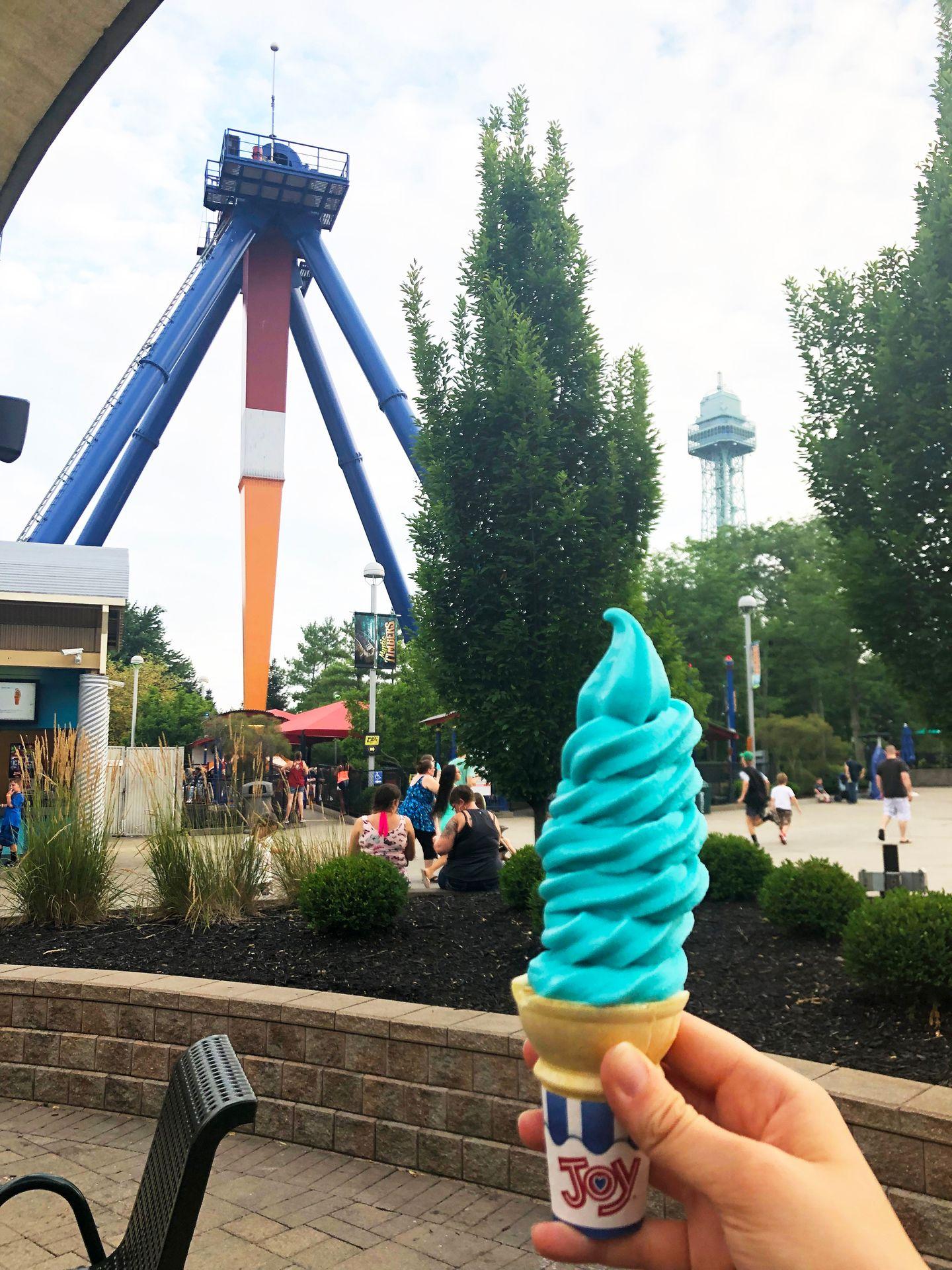 An ice cream cone with blue soft serve stacked high. There is an amusement park ride and the Kings Island Eiffel Tower in the background.