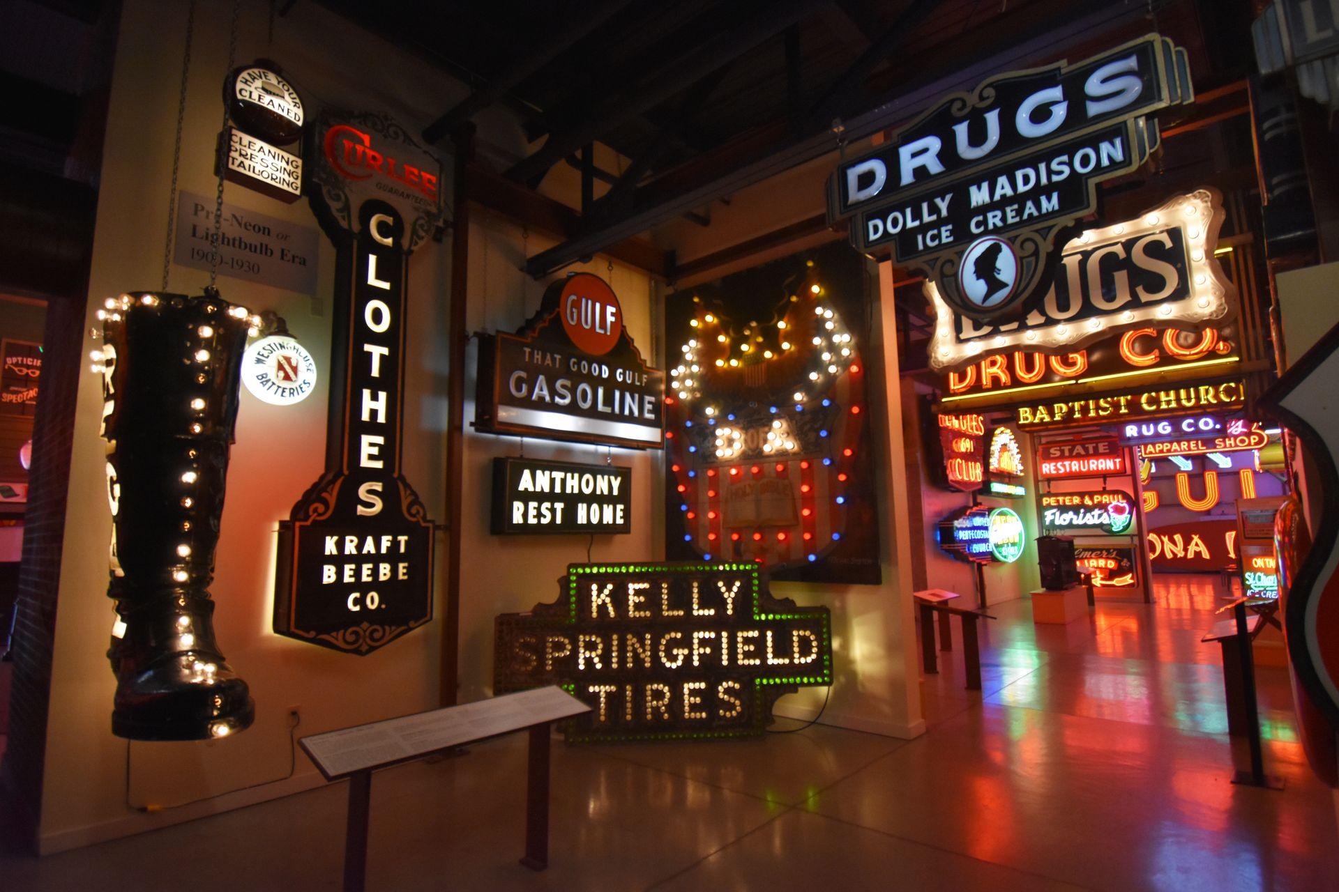 A photo of several neon signs inside the Sign Museum. There are signs for Drugs, Gasoline, Tires, clothes and a giant boot.