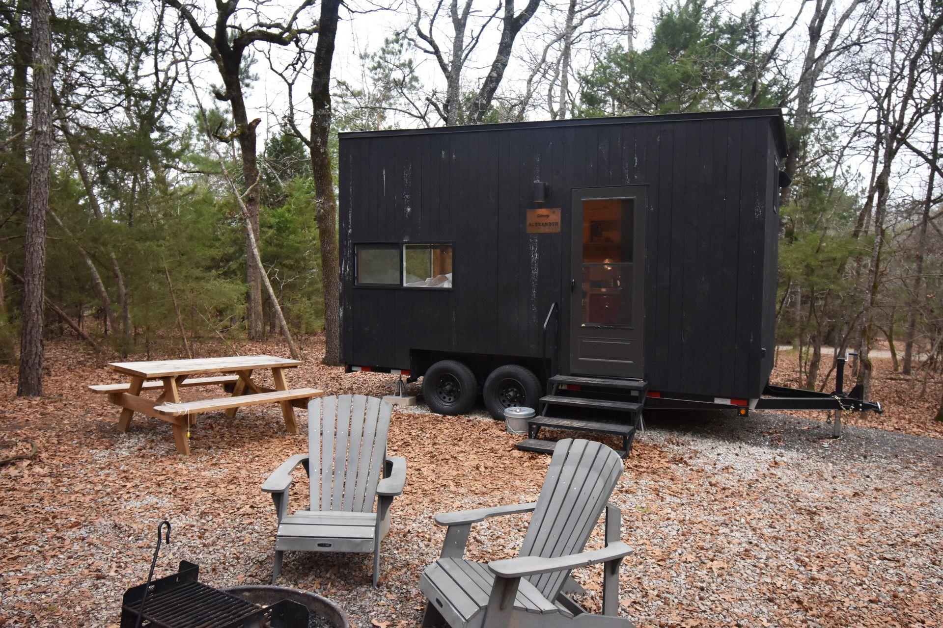 The exterior of the Piney Woods Getaway House. There is a tiny cabin, chairs surrounding a firepit and a picnic table.