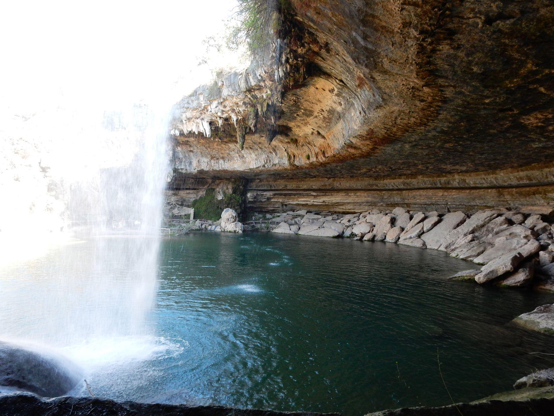 A pool of water with a cave overhanging above it. A waterfall flows down from the top of the cave and into the pool below.