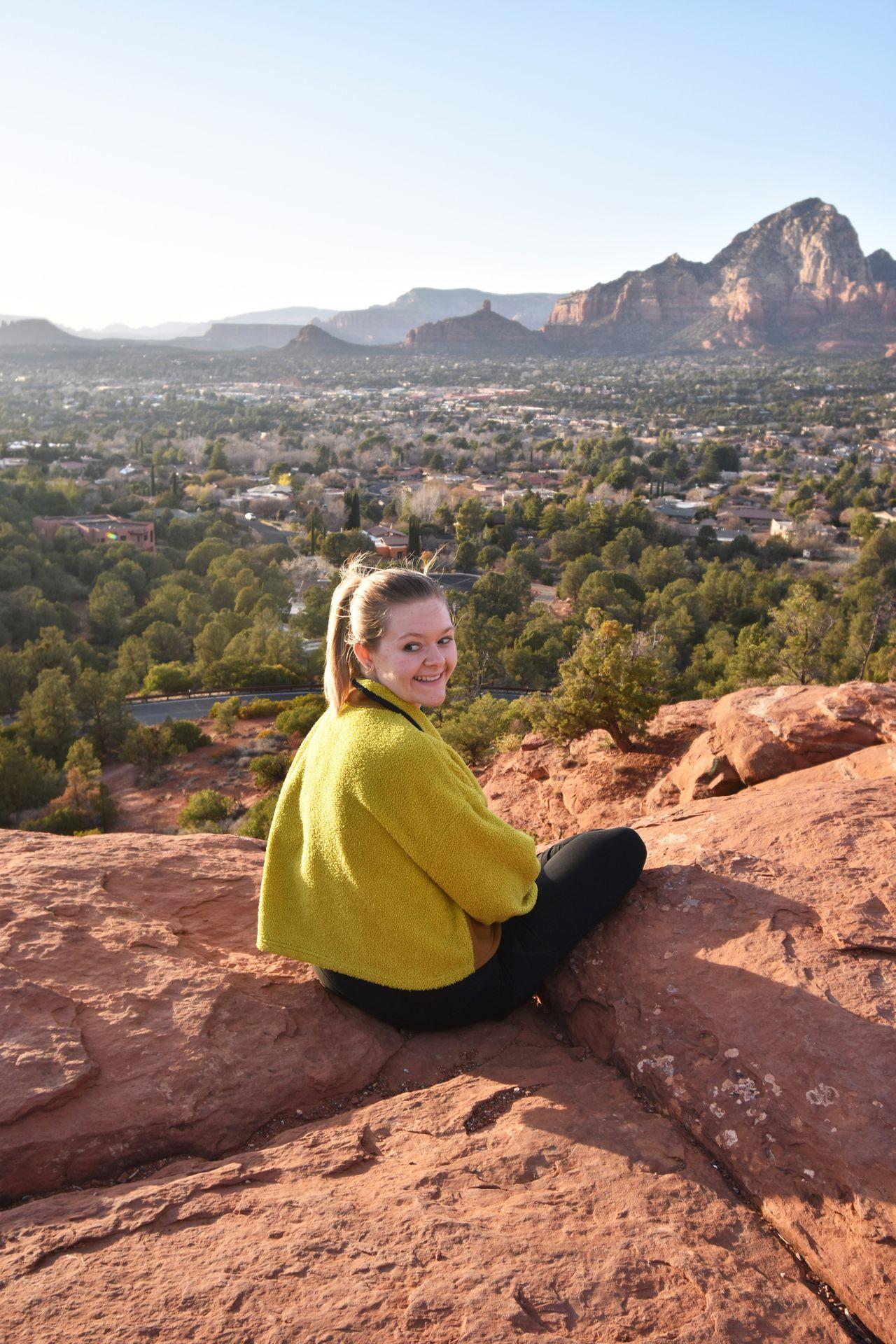 Lydia sitting on the edge of a rock wall on top of Airport Mesa. There are city and mountain views in the distance.