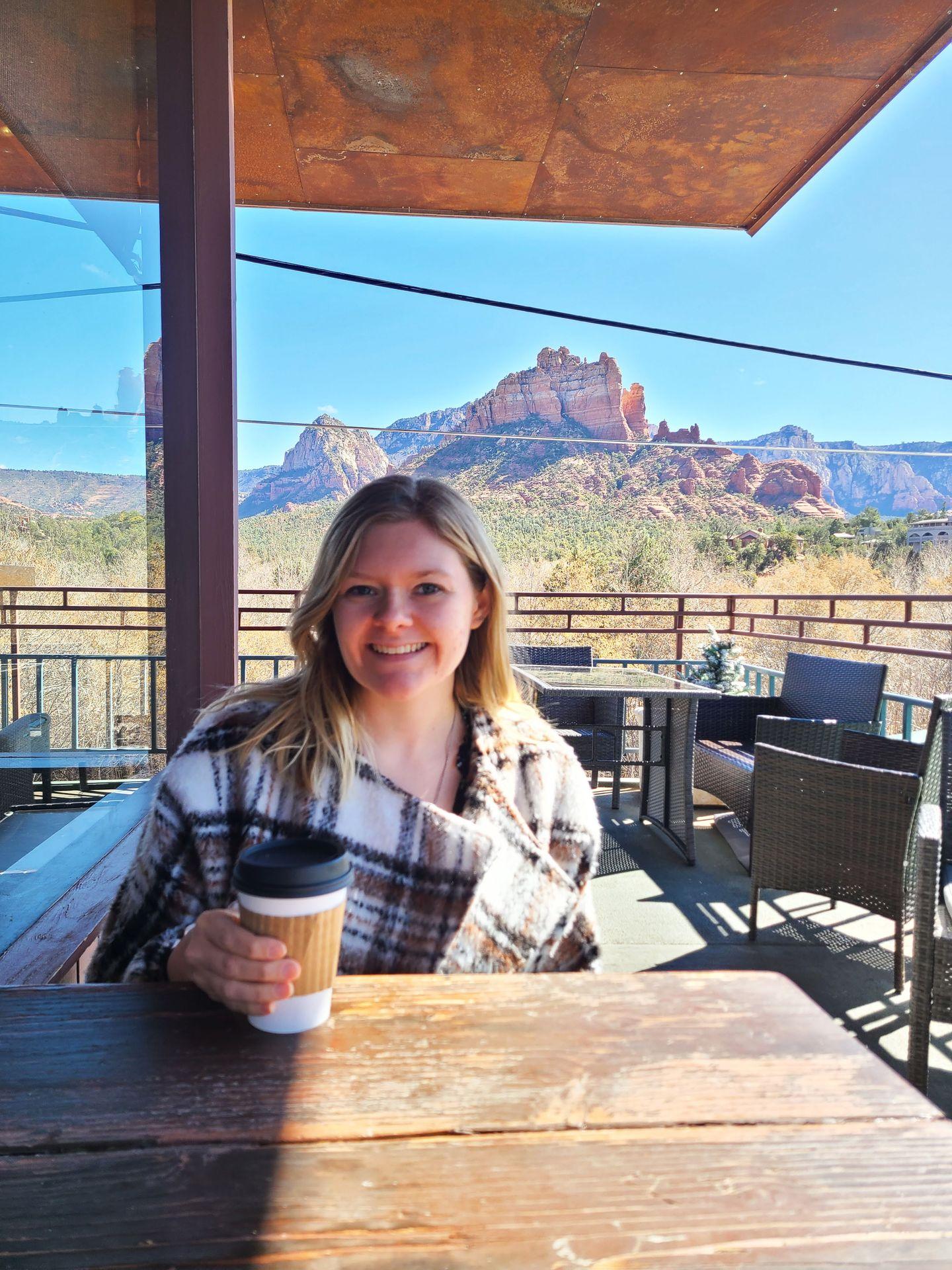 Lydia sitting at a table holding a coffee. There are orange rock views in the distance.