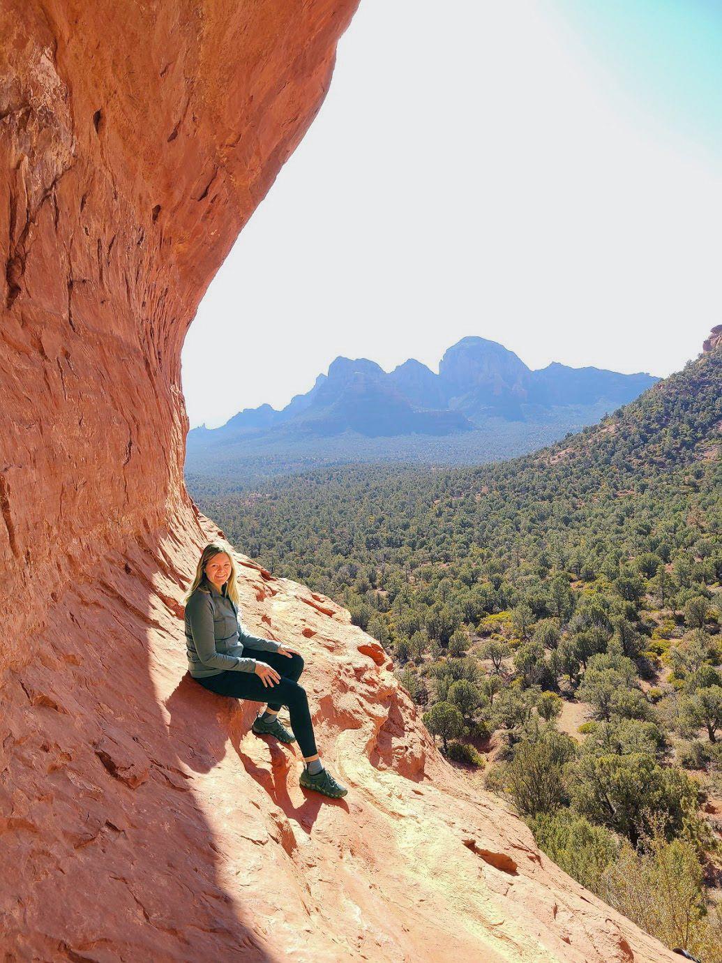 Lydia sitting on the side of the Birthing Cave with a view of mountains and trees behind her.