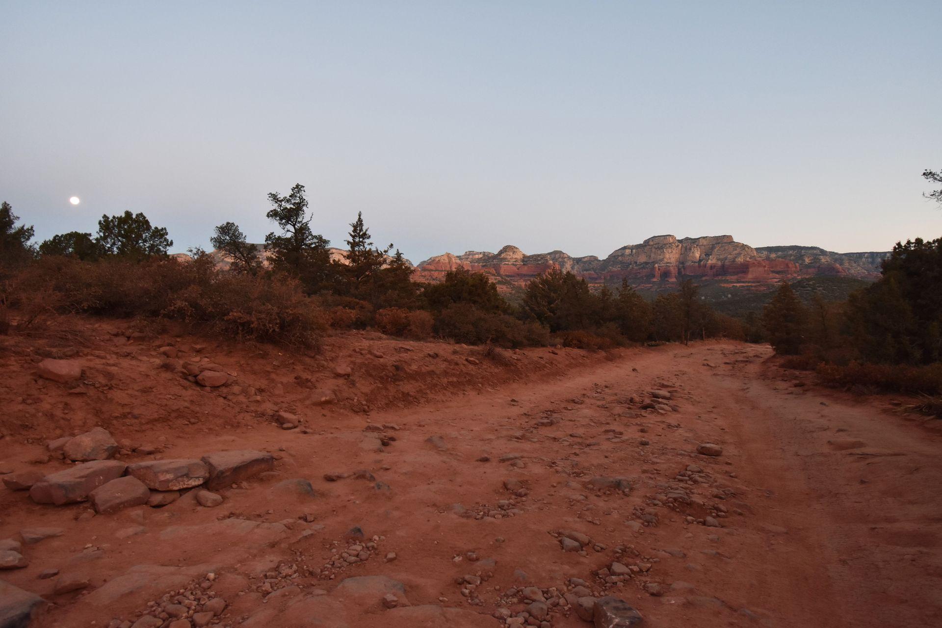 A wide, orange rock with rocks and dirt. There is a mountain in the distance and you can see the moon in the morning light.