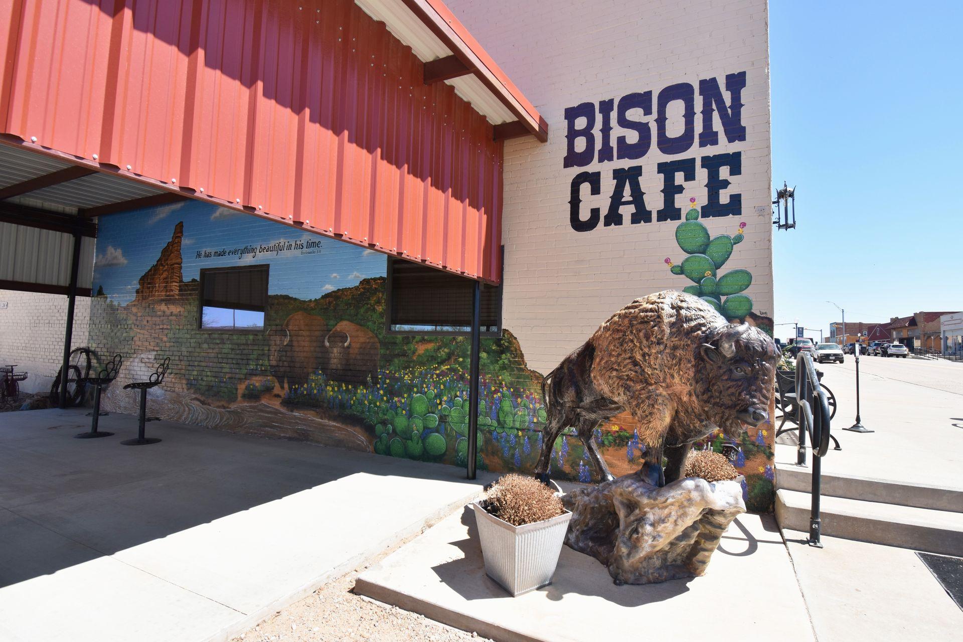 A bison statue in front of a buildng labeled "Bison Cafe." On the building is a mural painted with cacti and bison.