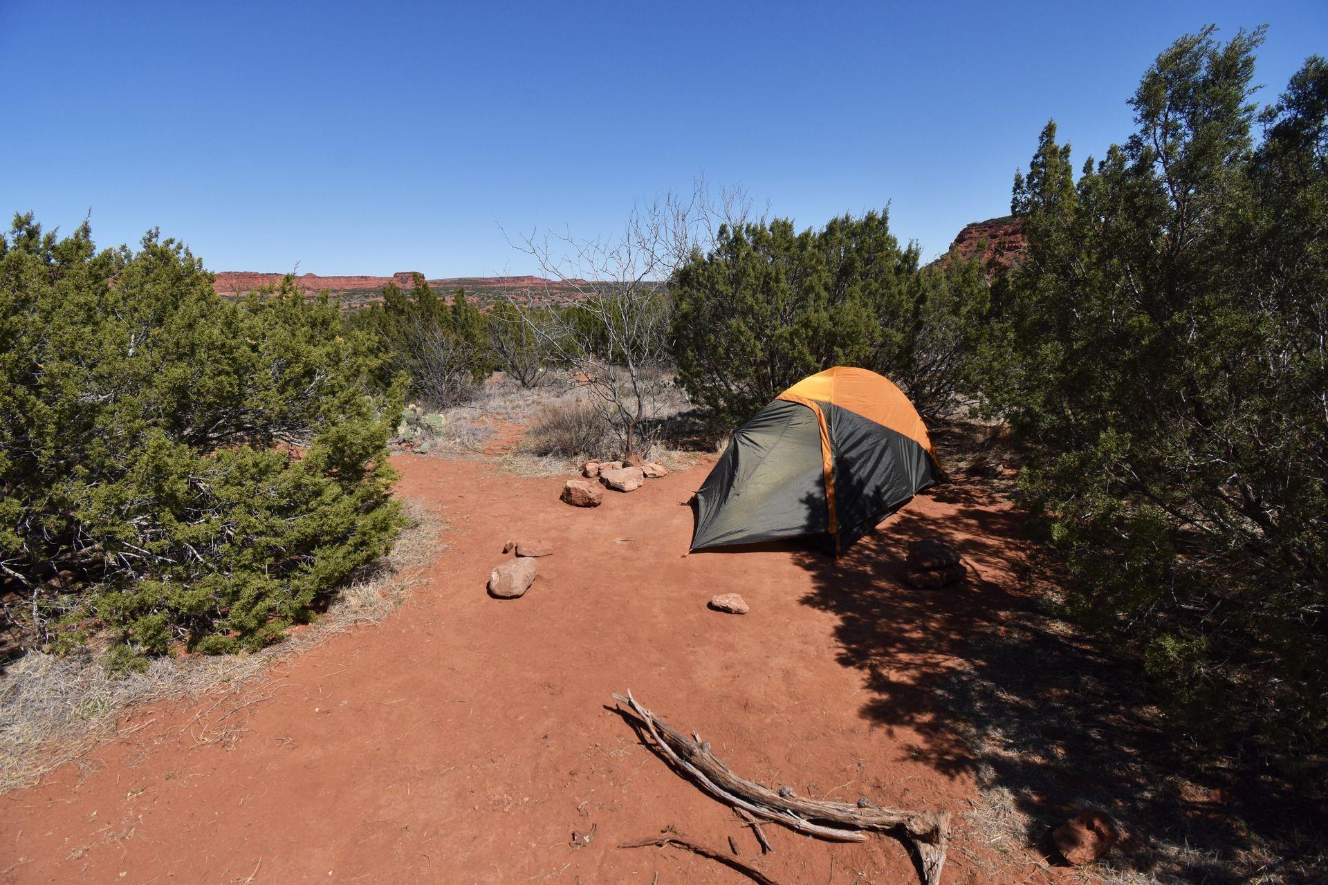 A tent set up on orange dirt and surrounded by some green trees.