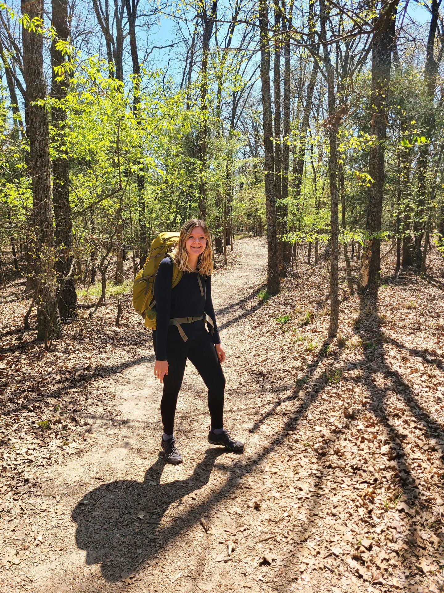Lydia wearing black and standing on a trail with a yellow backpack.