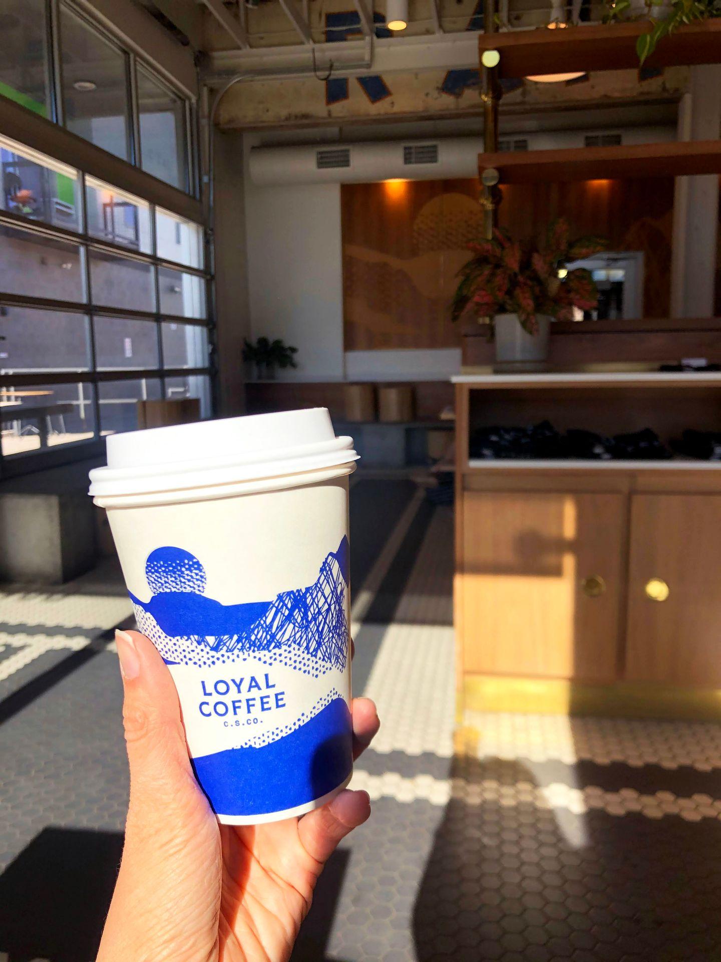 Holding up a to go cup of coffee inside of Loyal Coffee. The coffee mug has a blue and white landscape design.