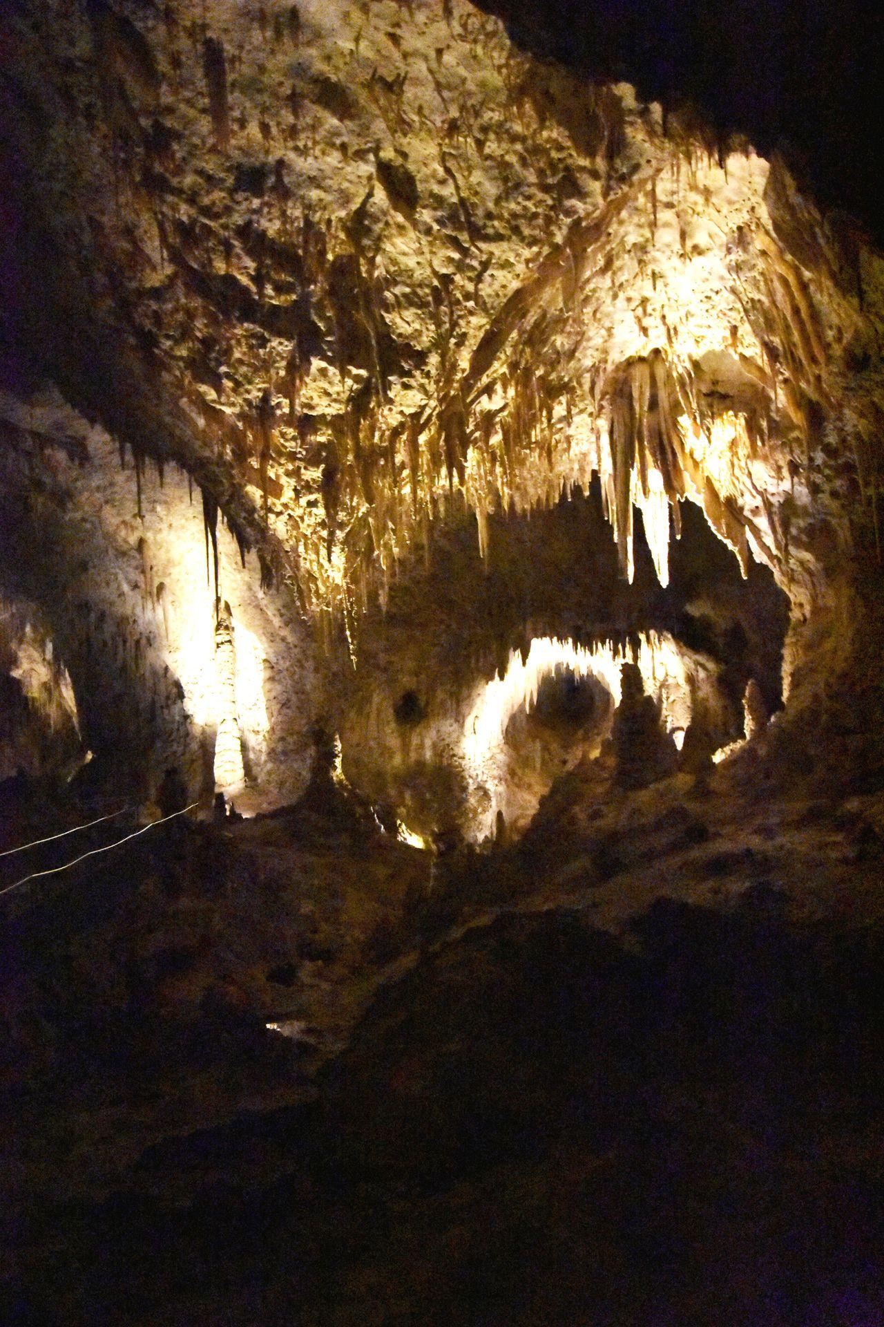 A dimly lit photo of the interior of the cave. There are several pointy stalactites and stalactites.