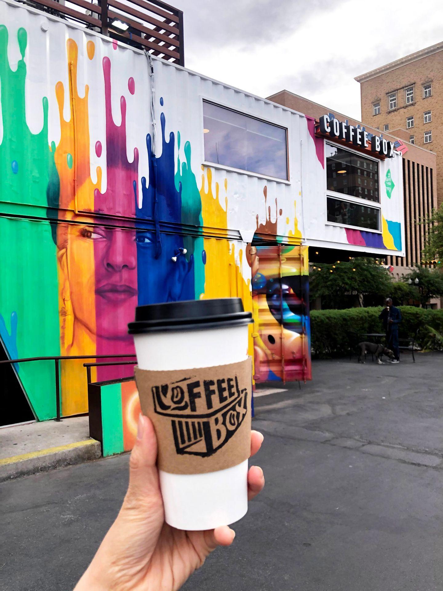 Holding up a coffee mug with a coffee shop made of shipping containers behind it. The building is painted with bright colors that look like a splash of paint.