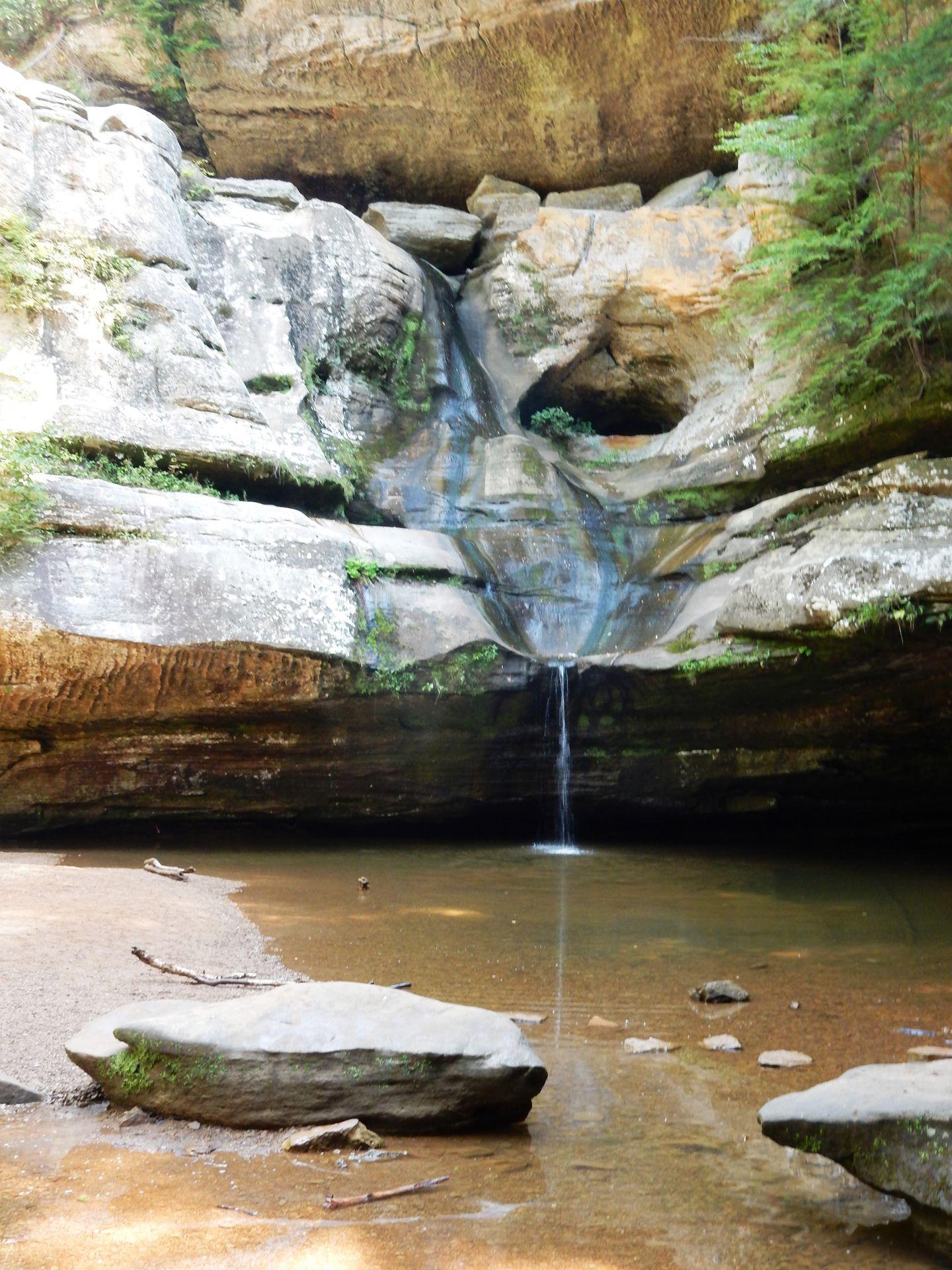 A view of Cedar Falls with a low water flow. Water trickles from a rock face into a pool of water.