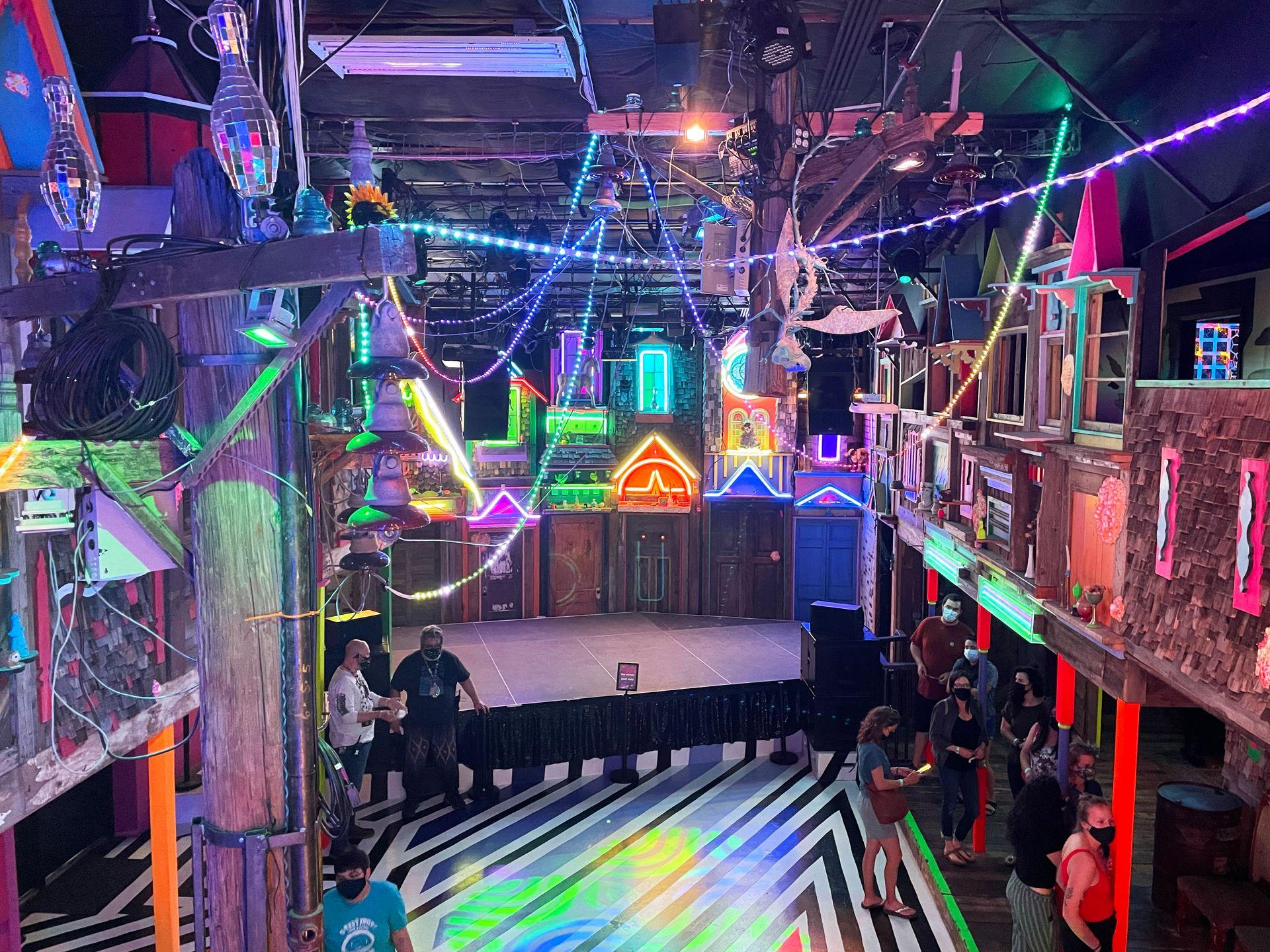 A room in Meow Wolf that is two stories tall and resembles a city. There are storefronts, bold stripes on the floor and colorful lights all over the room.