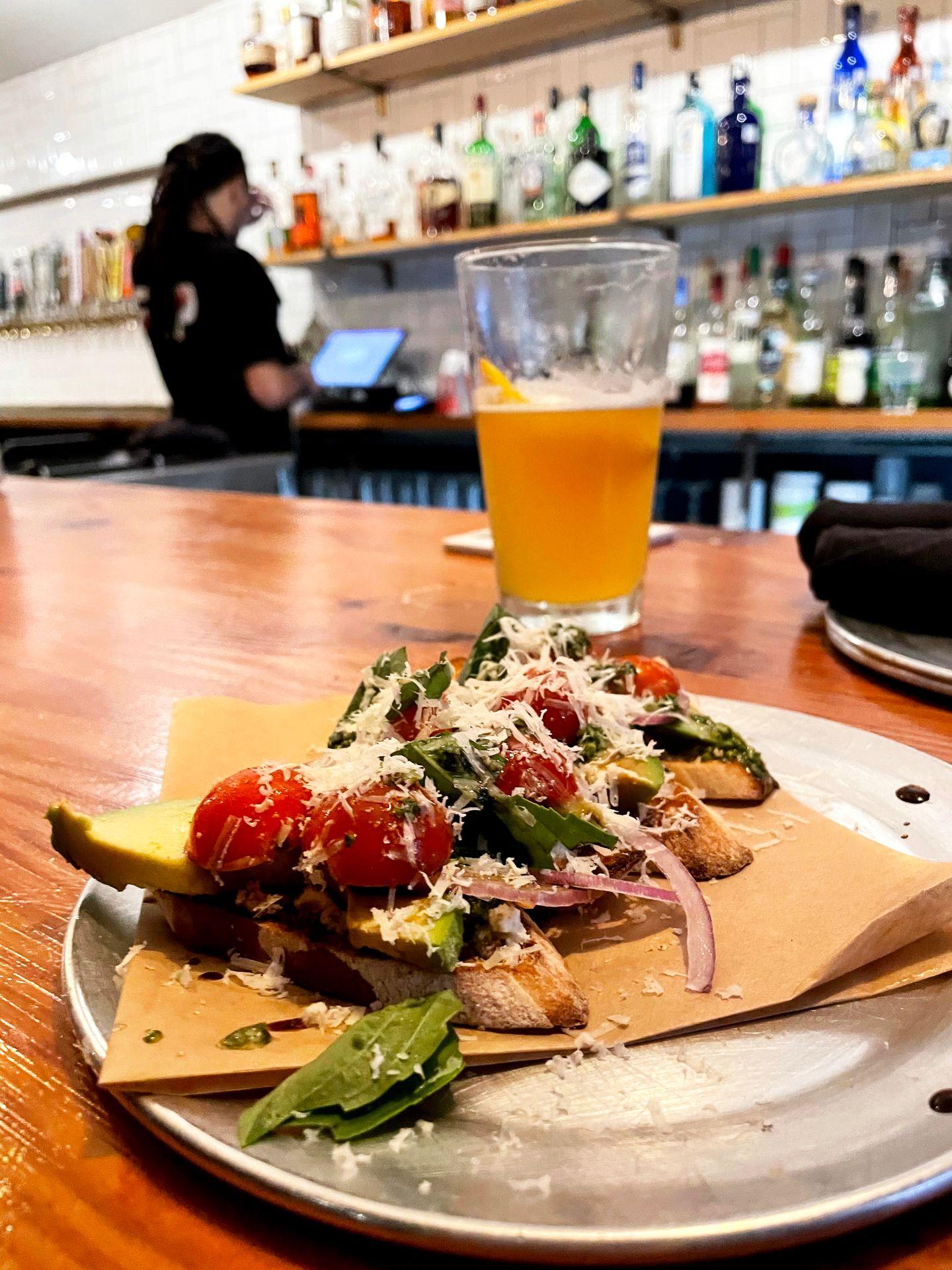A plate of bruschetta with a beer behind it at the bar at Community Pizza and Beer Garden.