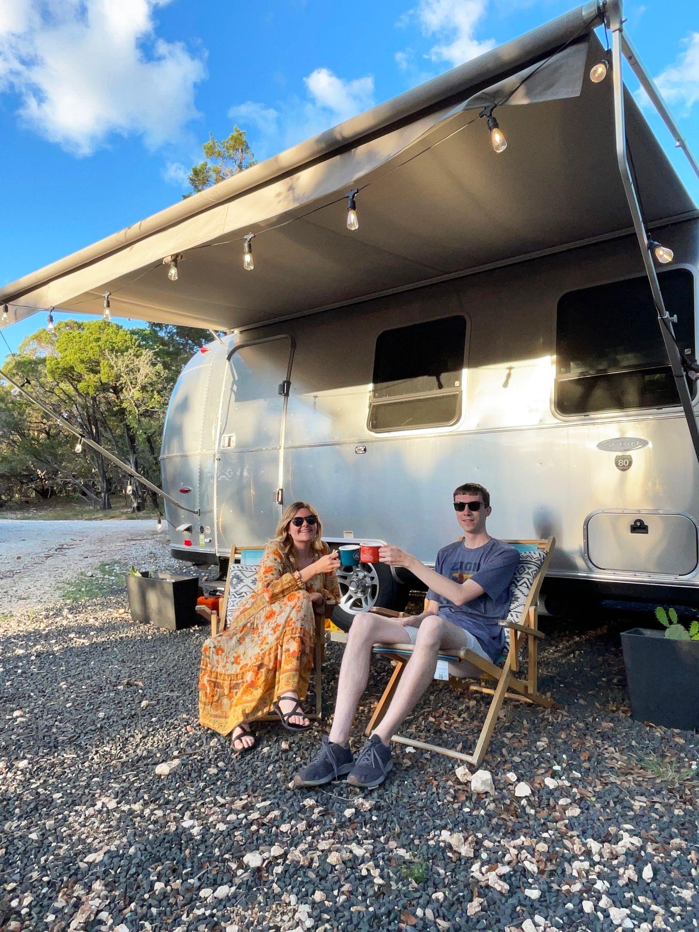 Lydia and Joe sitting in front of the trailer at Basecamp trailer Airbnb. They are tapping their mugs together.
