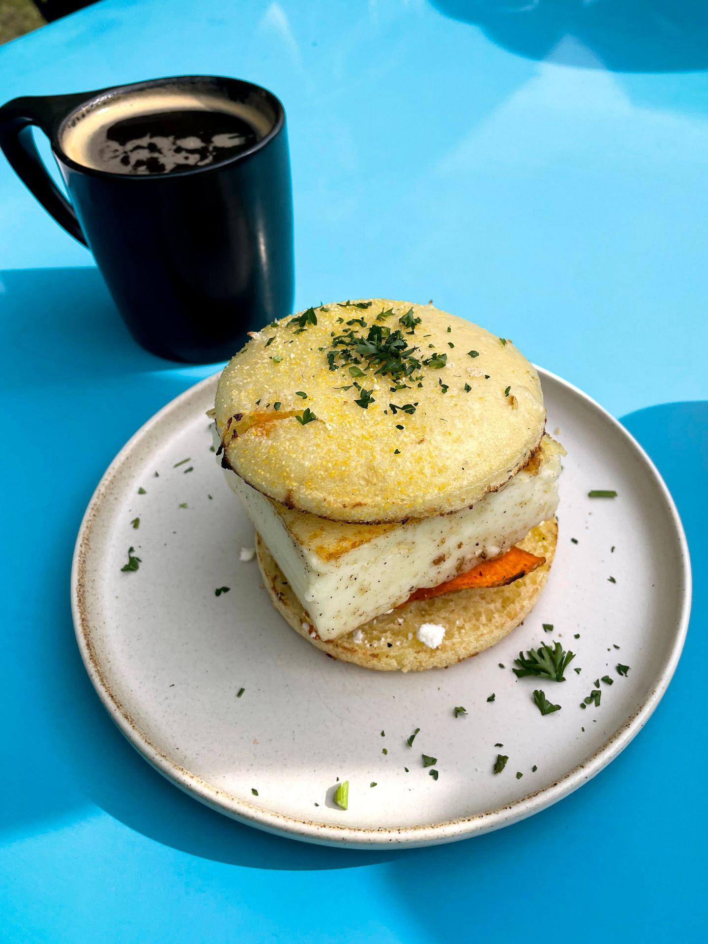 A breakfast sandwich with a square, souffled egg. It sits on a blue table next to a coffee mug.