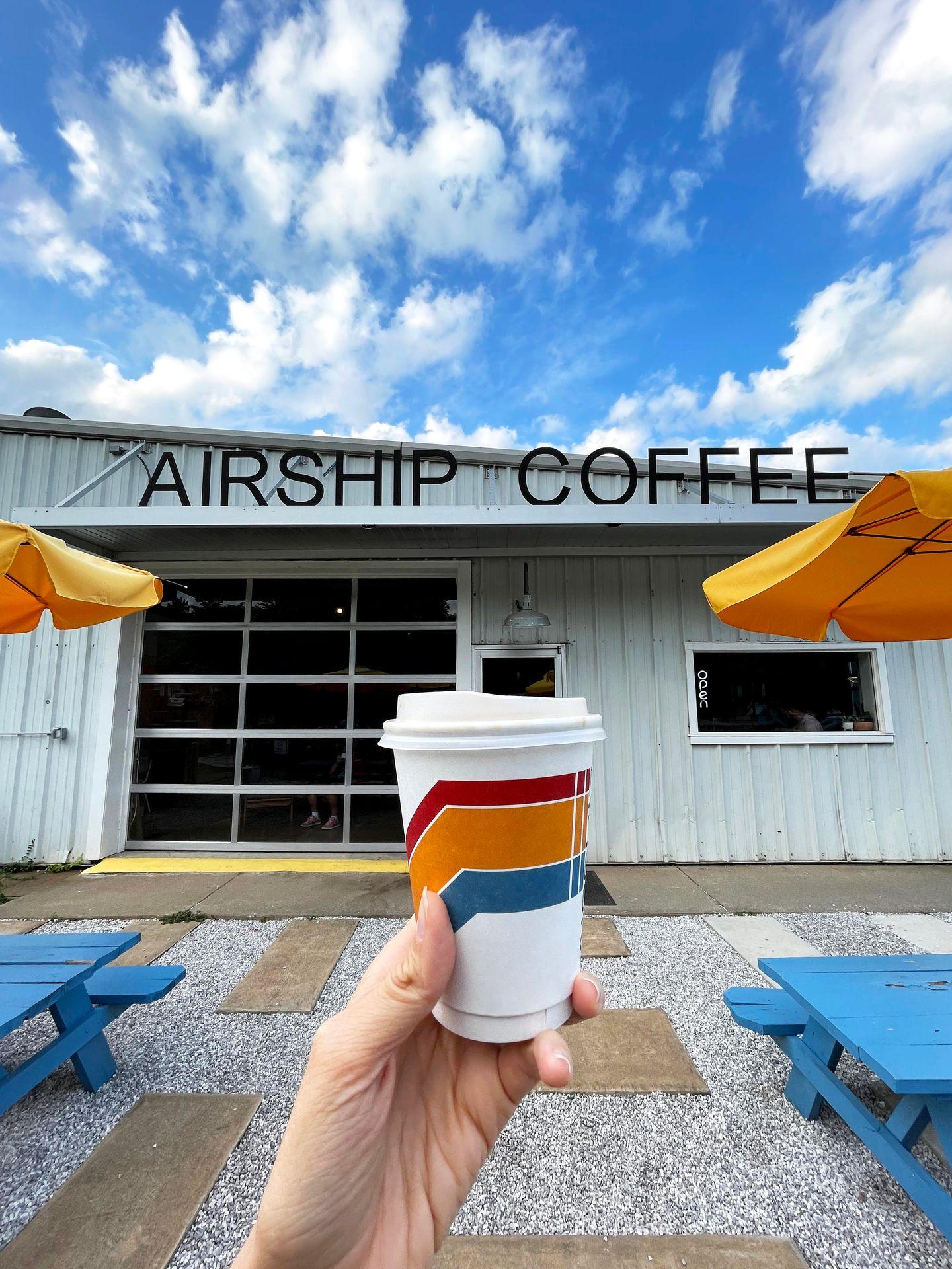 Holding up a to-go coffee mug that has a red, orange and blue stripe design. In the background is the exterior of Airship Coffee.