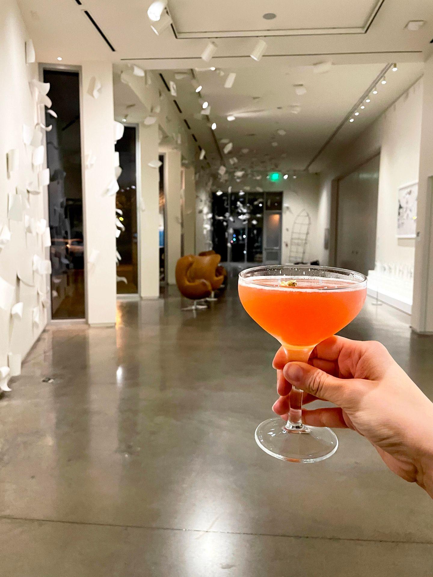Holding up an orange cocktail with an art gallery in the background.