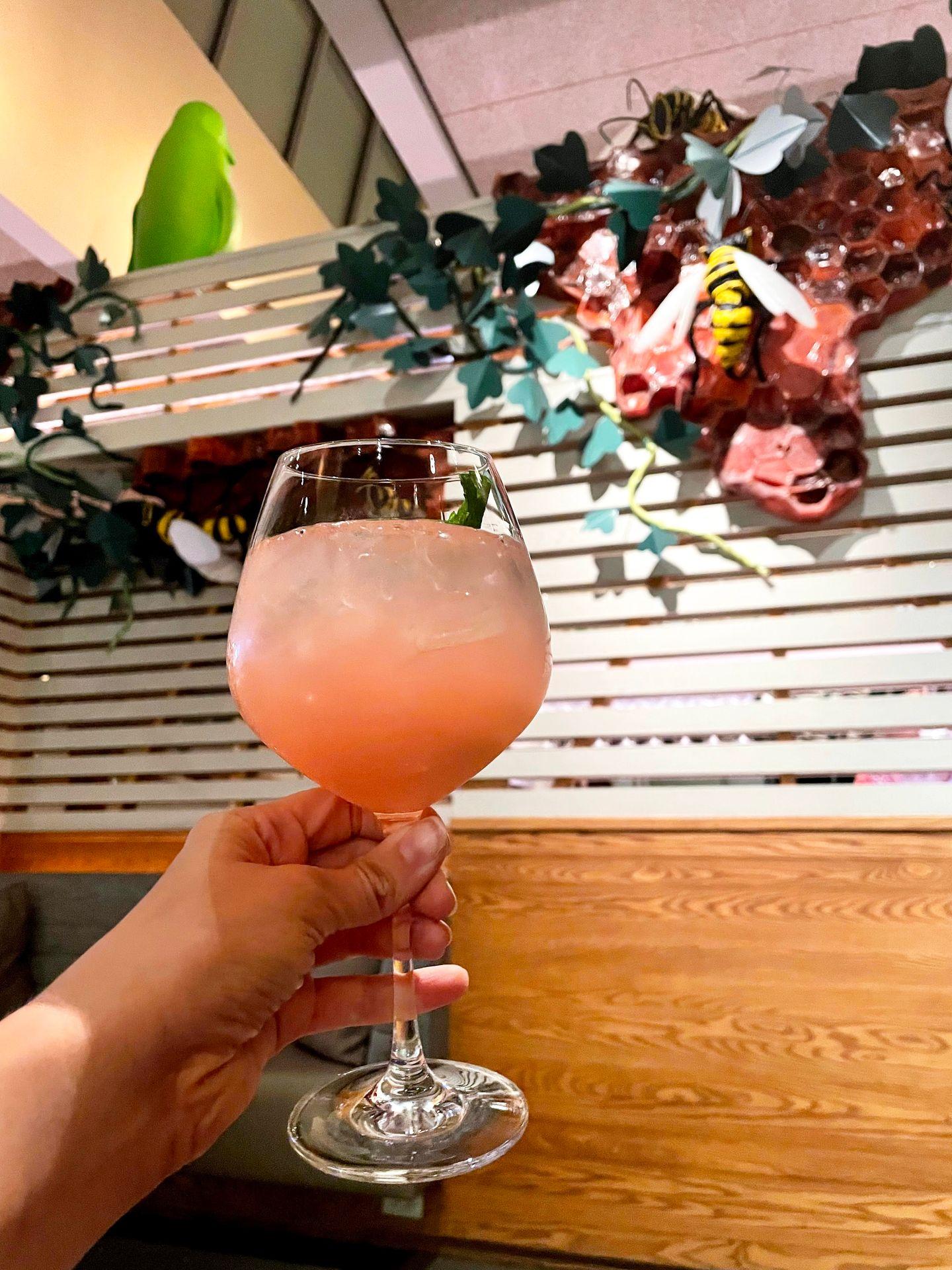 Holding up a pink cocktail inside of The Hive. There is artwork that resembles a beehive and plants hanging from the wall and a green penguin up high facing the other way.