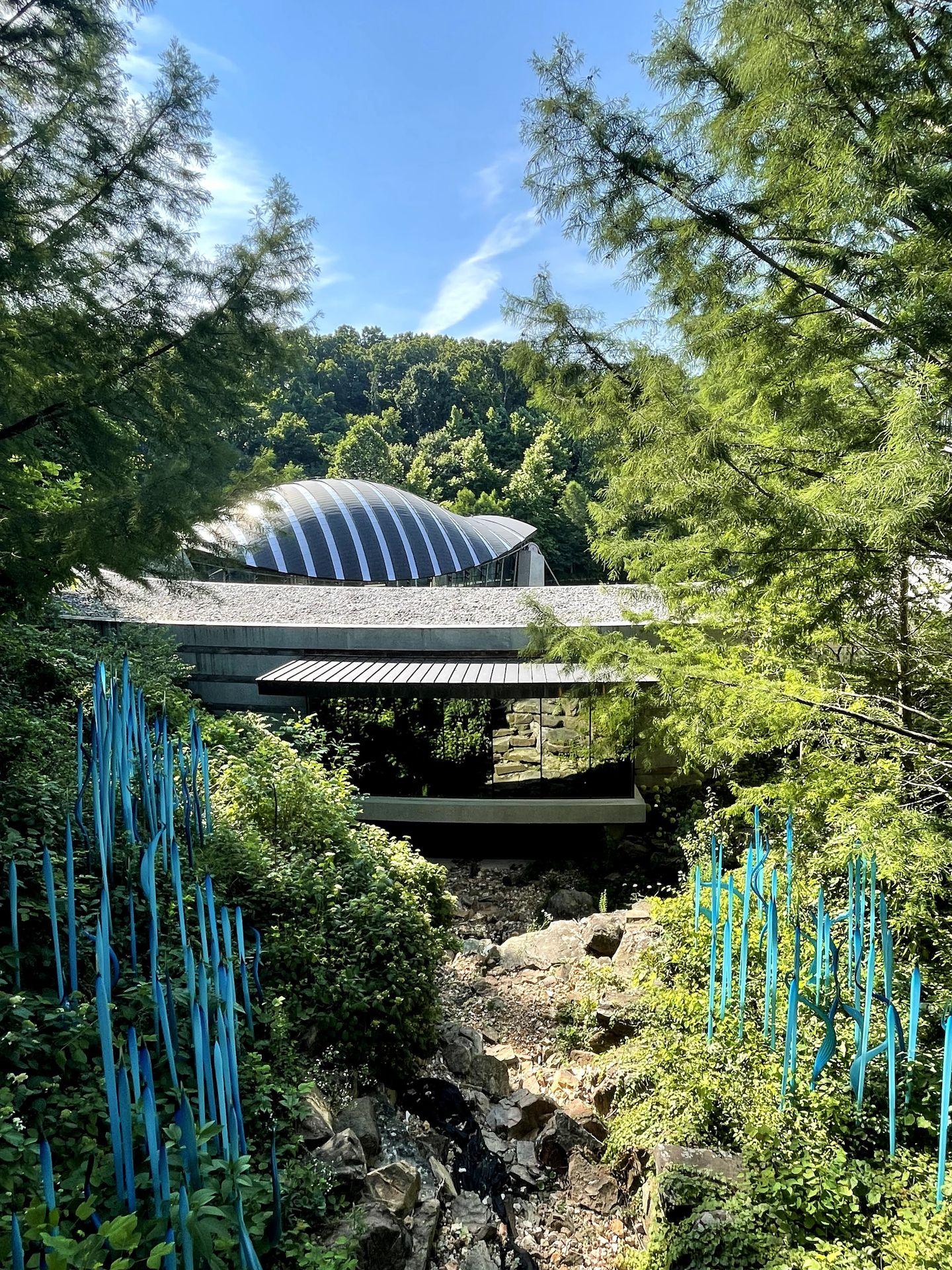The exterior of the Crystal Bridges from a distance. There is a curved building, trees and some blue sculptures outside.