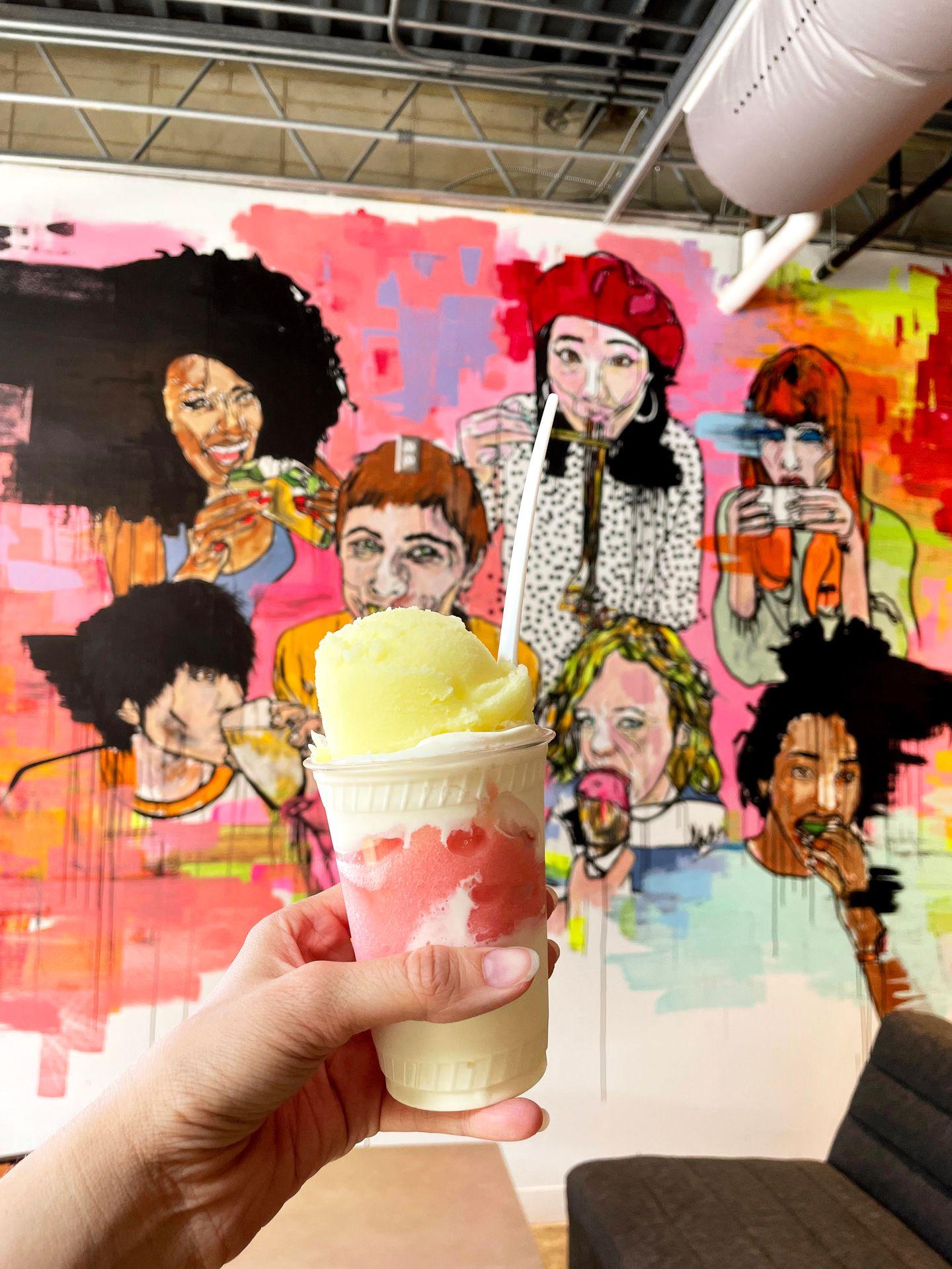 A cup with custard layered with pink and yellow Italian ice. There is a colorful mural in the background.