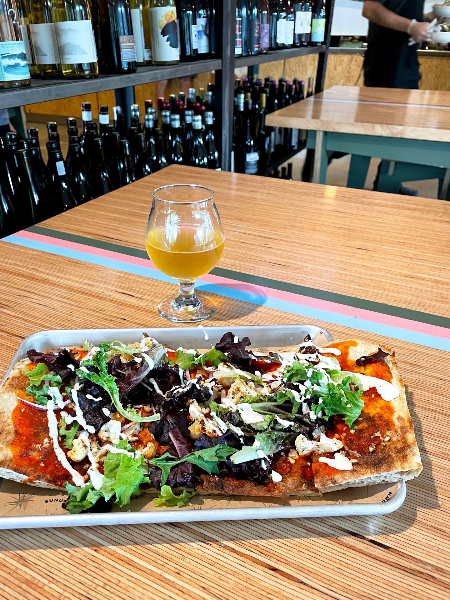 A flat bread pizza topped with lettuce and cheese. A glass of beer sits on the table next to it. The table is light wood with three stripes: one pink, one green, one blue.