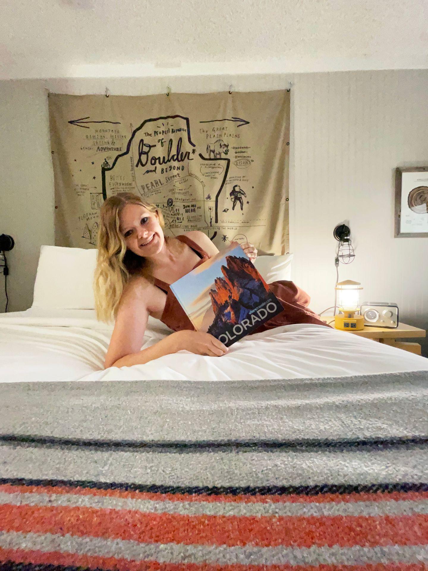 Lydia on a bed holding a Colorado book. There is an artistic map of Boulder on the wall in the background.