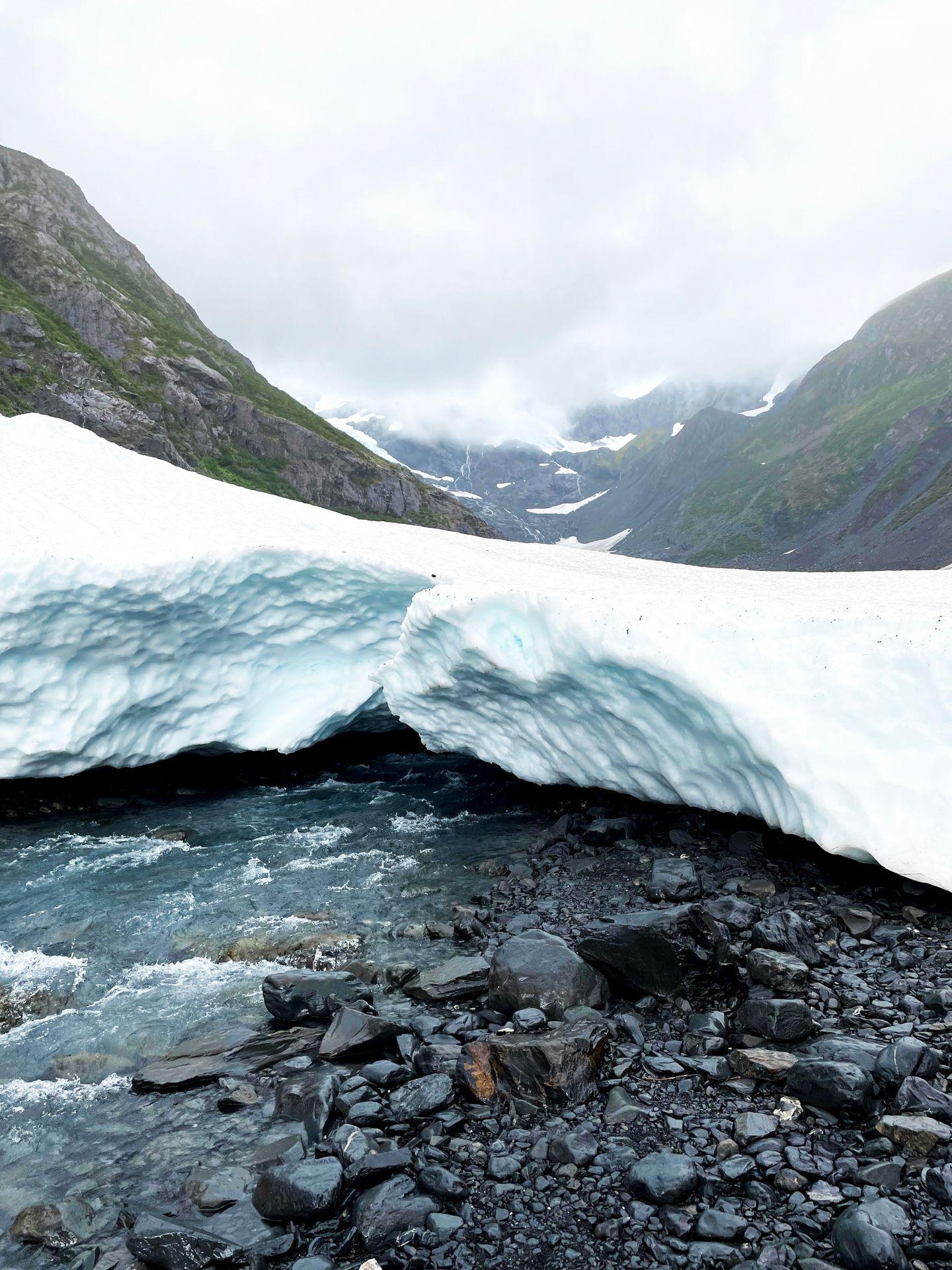 A small glacier with a river flowing under it and mountains in the background.