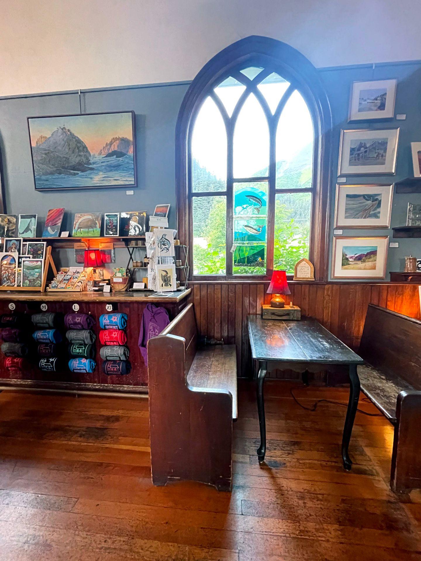 Inside of Resurrect Art Coffee House. There is a window that looks like it's from a church, gifts for sale and a table with pews as benches.