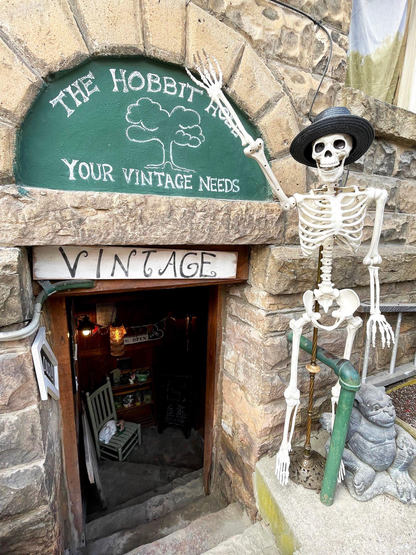 The exterior of the Hobbit Hole shop. There is a skeleton outside of the door and the steps lead down below street level.