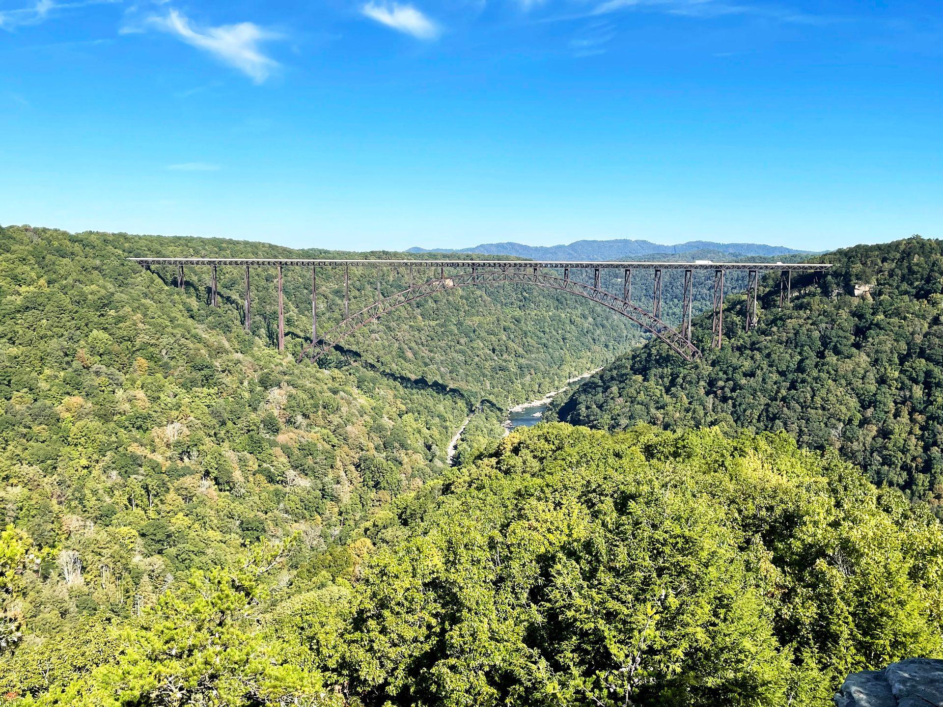 A view of the New River Gorge Bridge from the end of the Long Point Trail. It is surrounded by green trees.