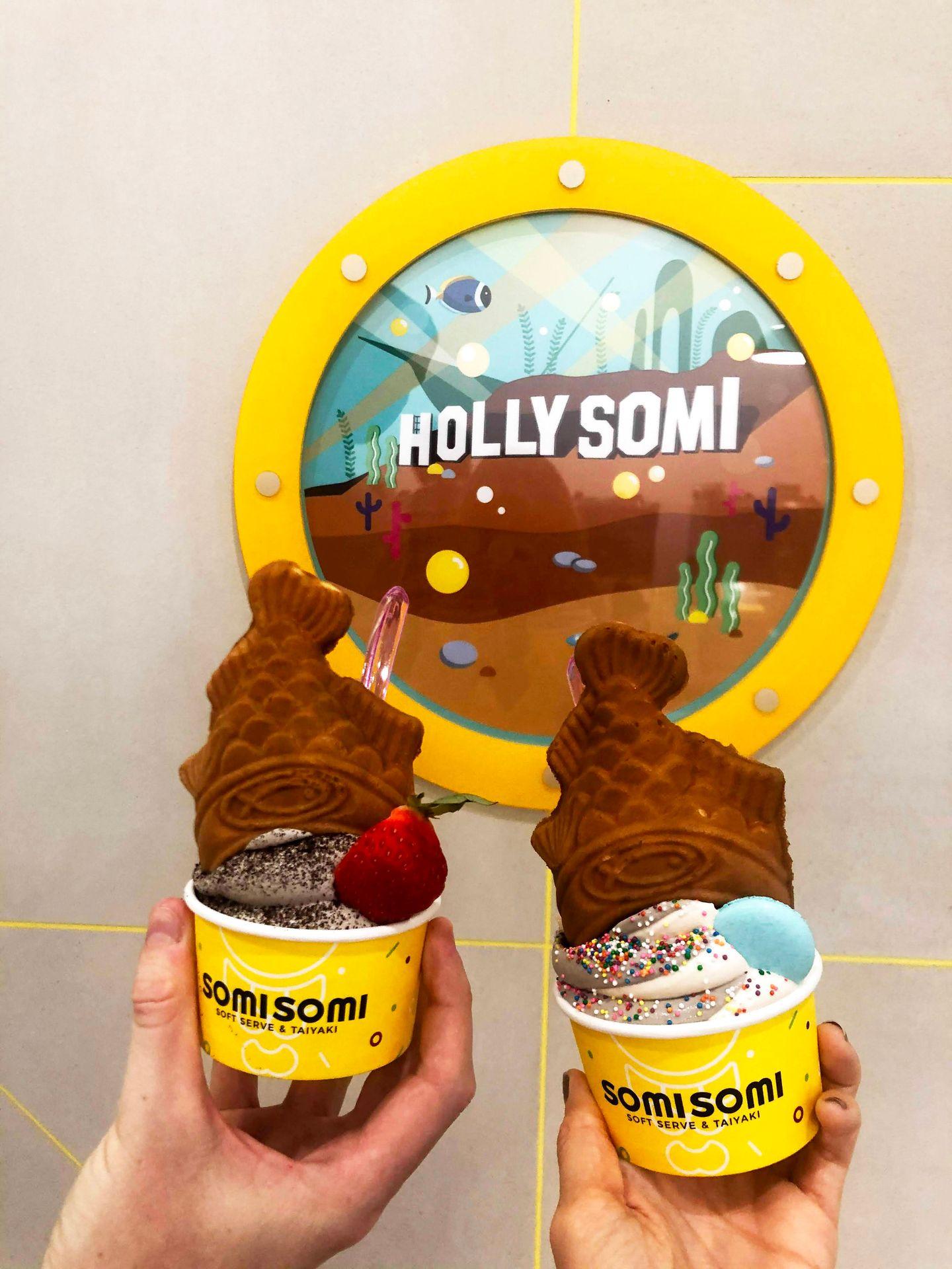 Two hands holding cups of cream with a taiyaki cone shaped like a fish on top. A sign reads "Hollysomi" in the background.