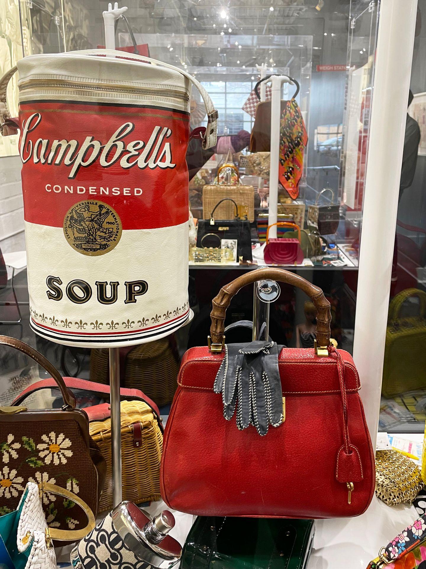 A close up view of a Campbell's soup purse and a red purse with some gloves draped over it.