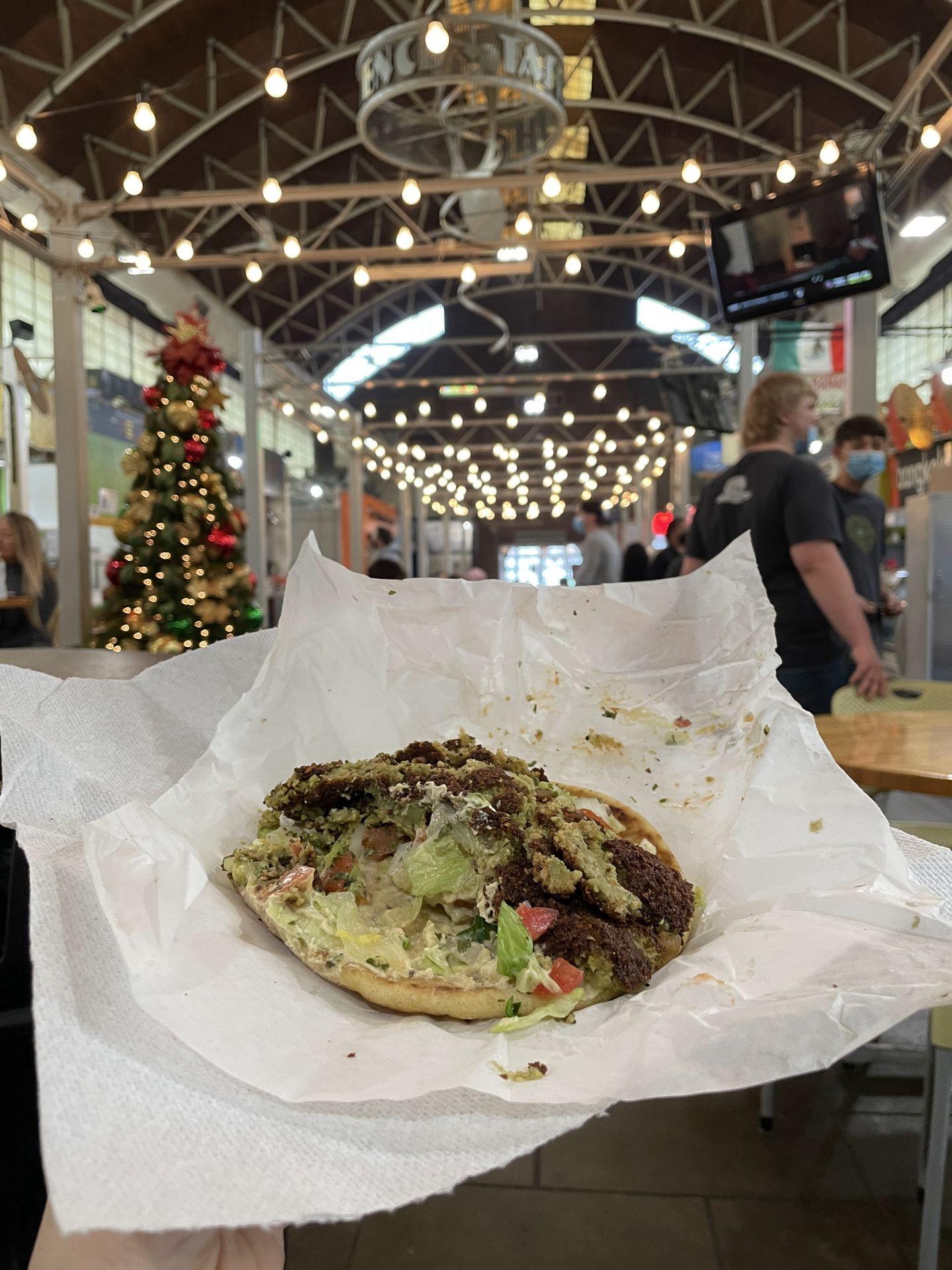 A falafel gyro being held up in the Little Rock Central Market. There is falafel, lettuce and tomato on pita bread.