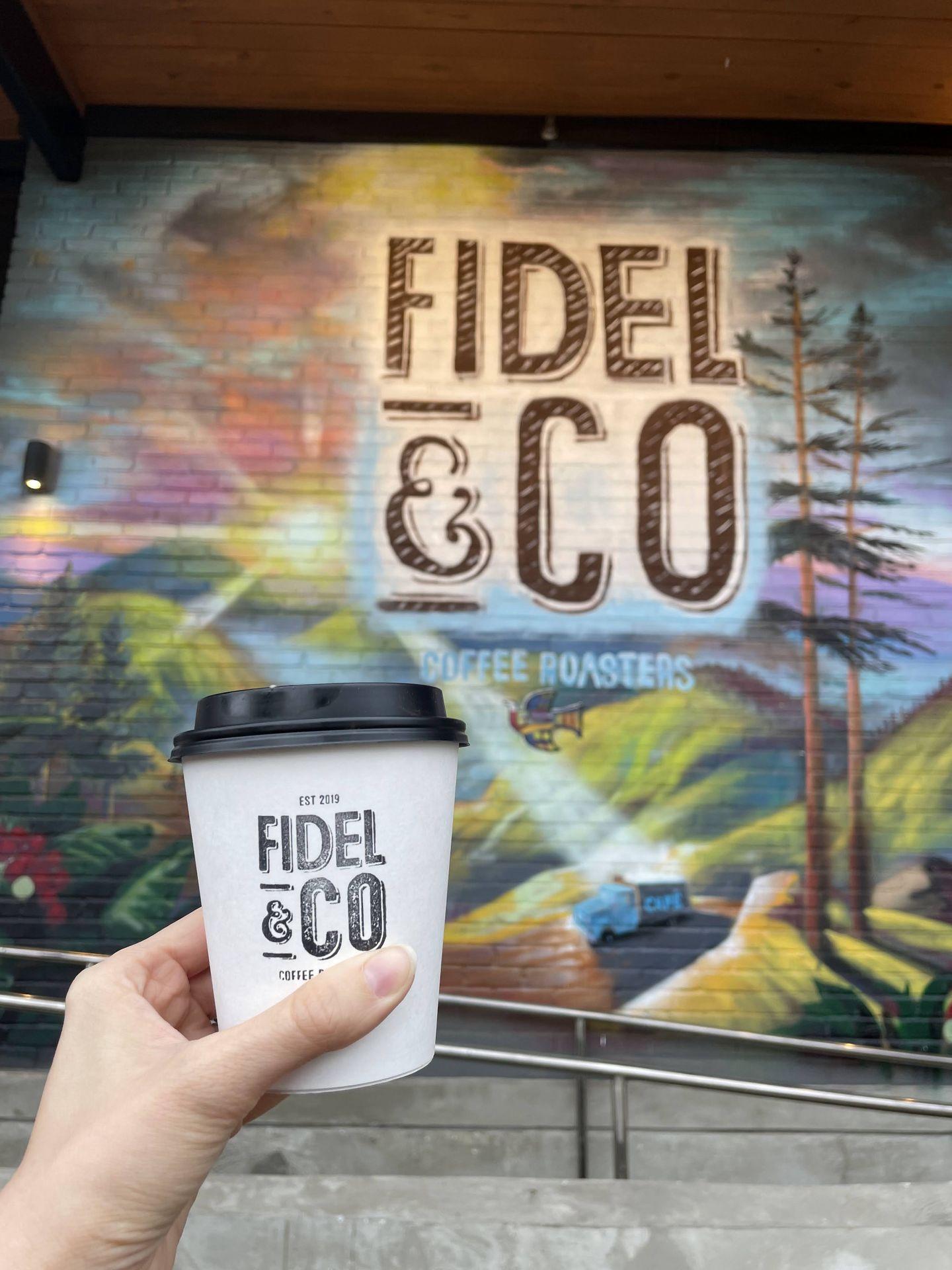 Holding up a coffee cup in front of a mural. The mural has green mountains, greens and a colorful sky, with the words 'Fidel & Co' in the middle.