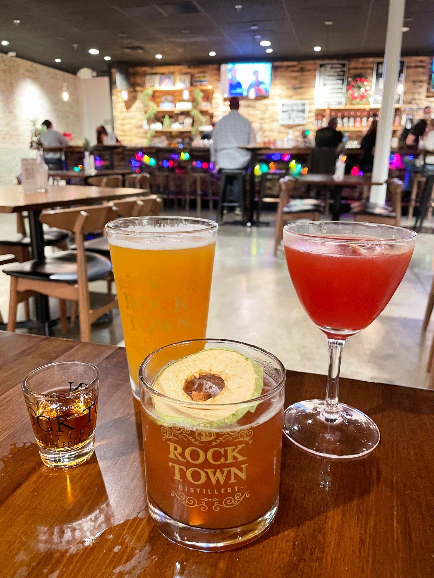 Four drinks on a table inside of Rock Town Distillery. There is a beer, a red cocktail, a short cocktail with a garnish and a shot glass.