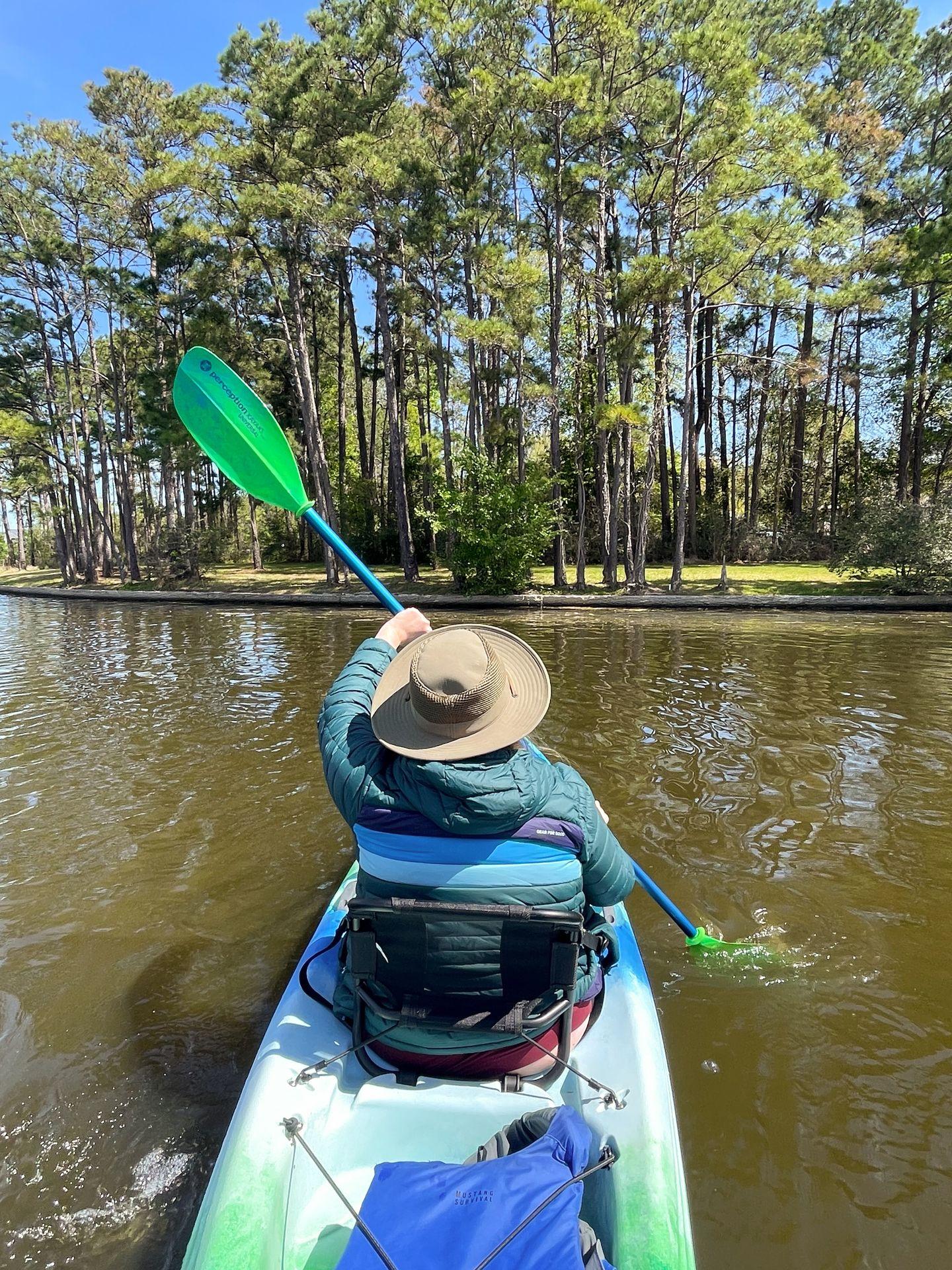 Lydia kayaking away from the camera and towards tall trees on Lake Woodlands. She wears a puffer jacket and a hat.