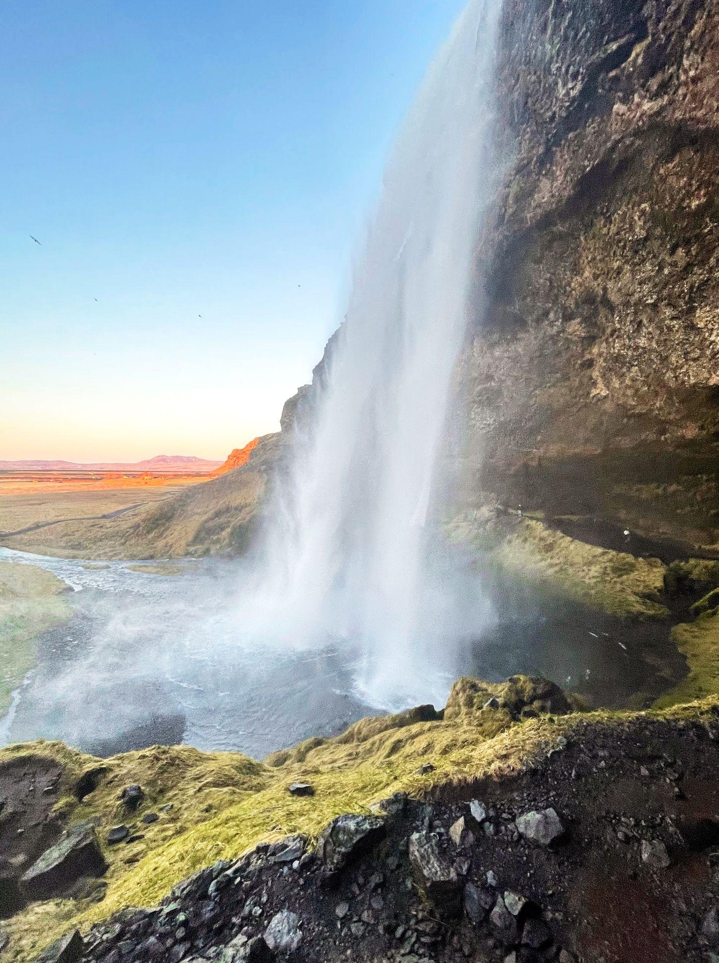 The Seljalandsfoss waterfall in Iceland. It flows down over the walls of a canyon. The sun is setting in the distance.