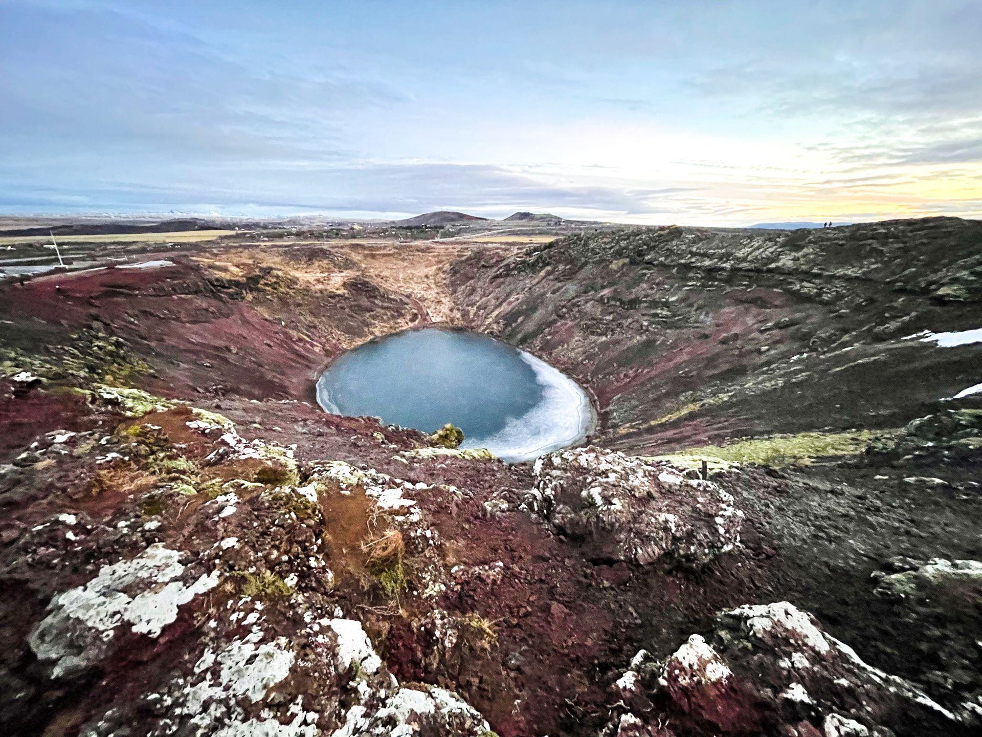 Looking down at a round crater. The crater has blue water and the cliff faces have a red color.