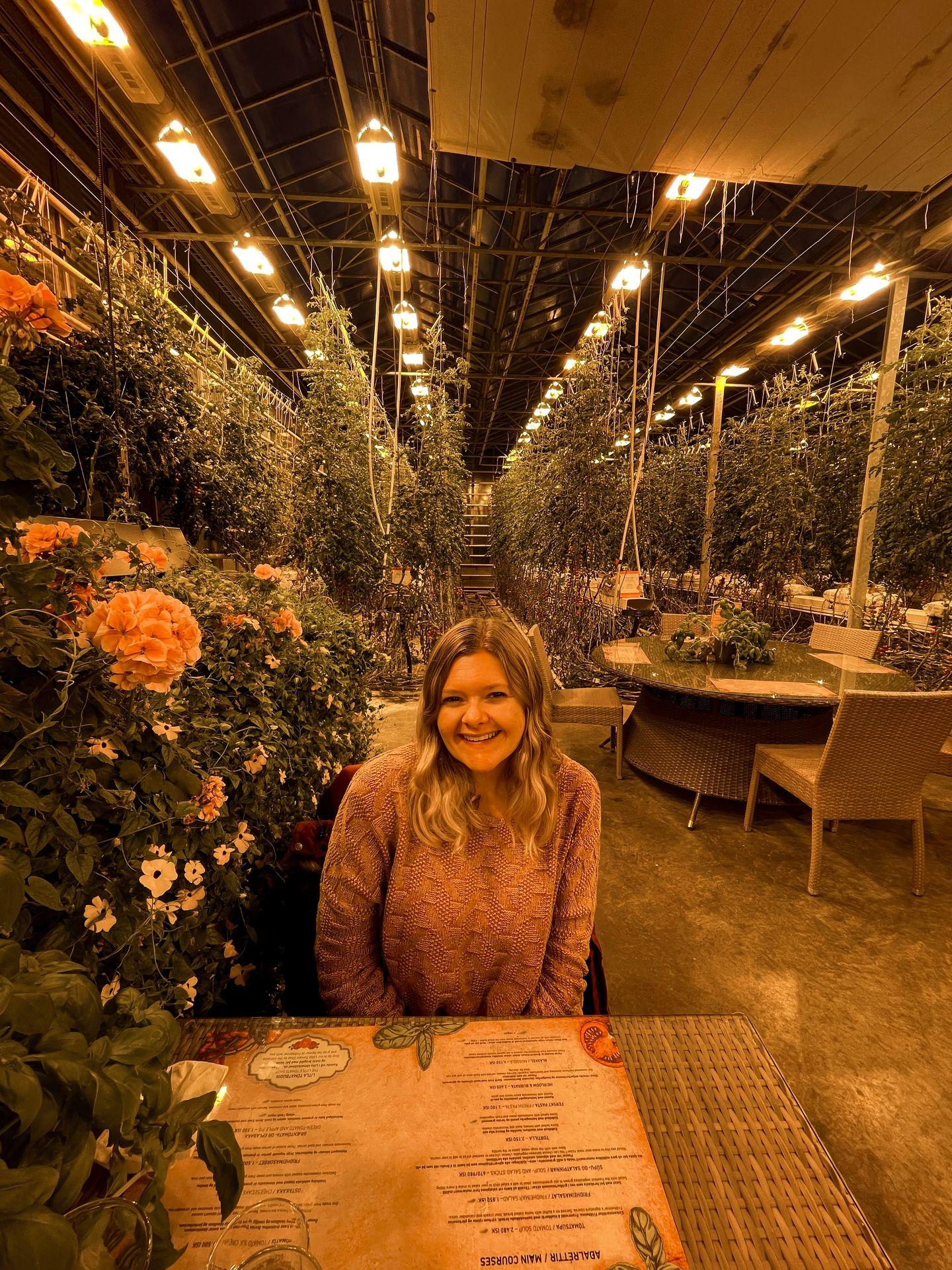 Lydia sitting at a table inside of Friðheimar. She is surrounded by green vines with flowers.