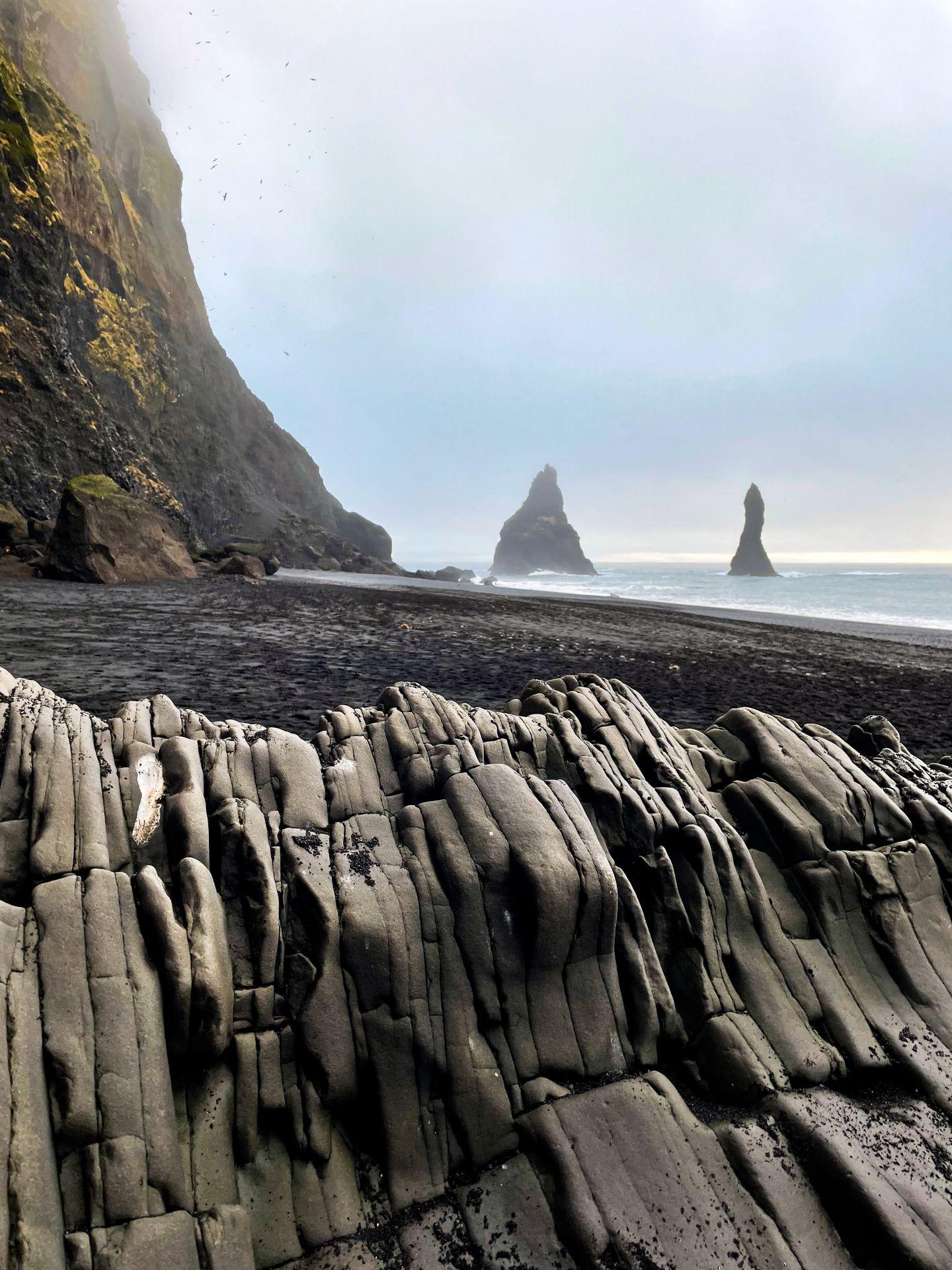 Textured rocks with a black sand beach behind them. There are two large, triangle-shaped rock rising out of the water in the distance.