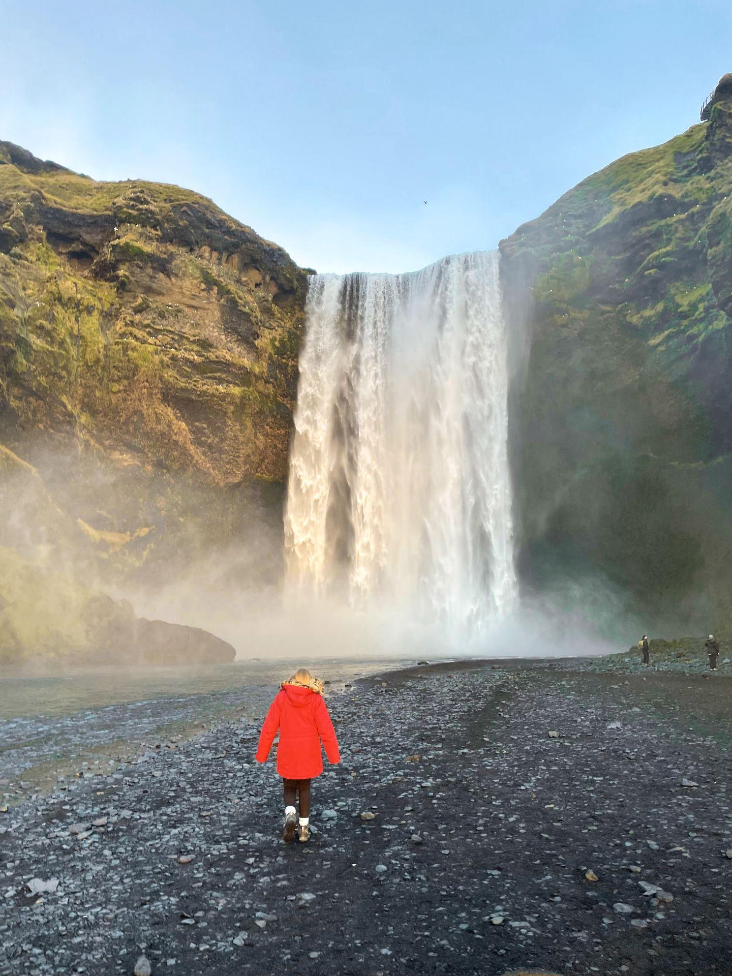 Lydia wearing a red coat and walking towards the towering Skógafoss