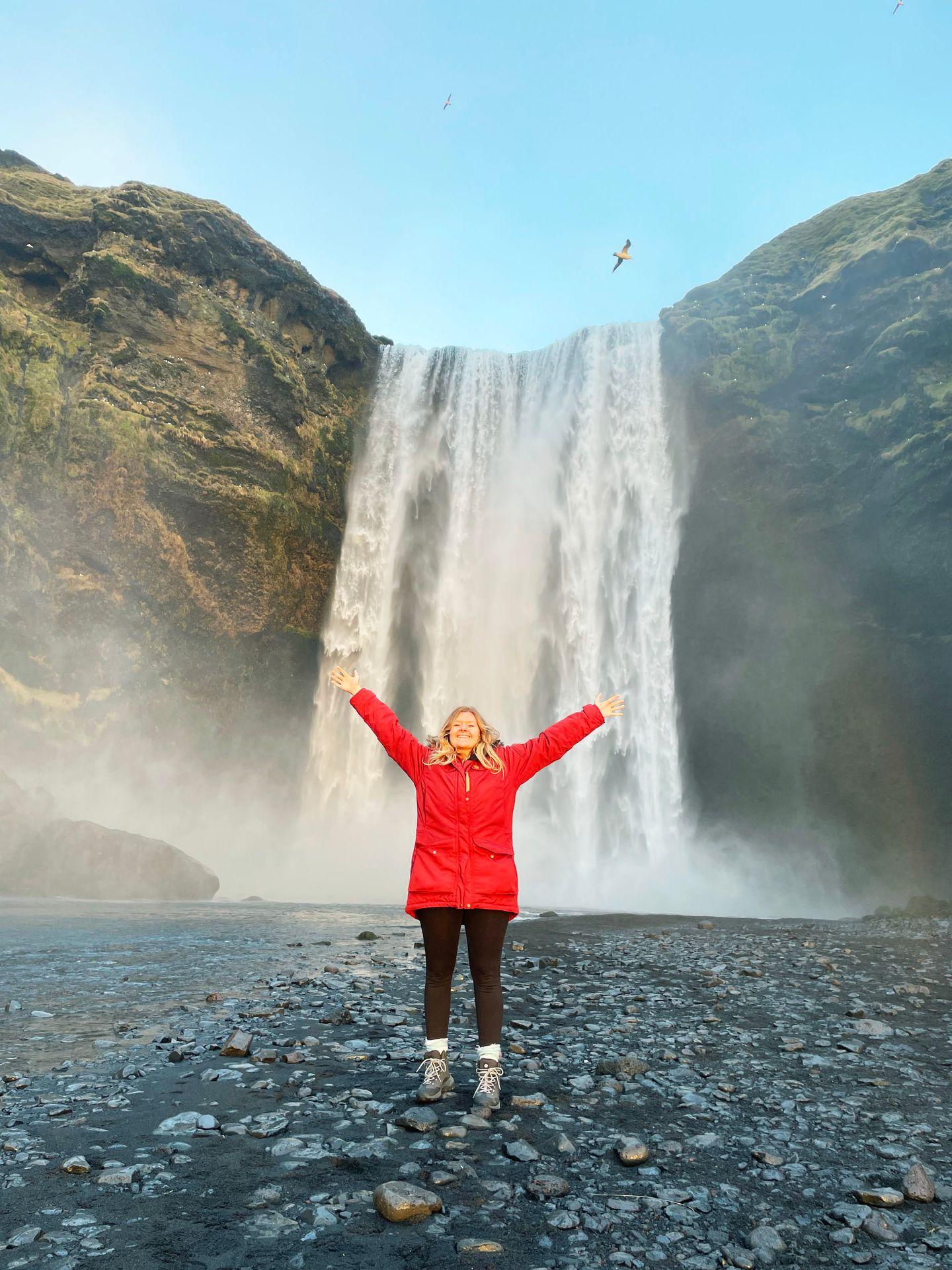 Lydia standing in front of a huge waterfall with her hands in the air.